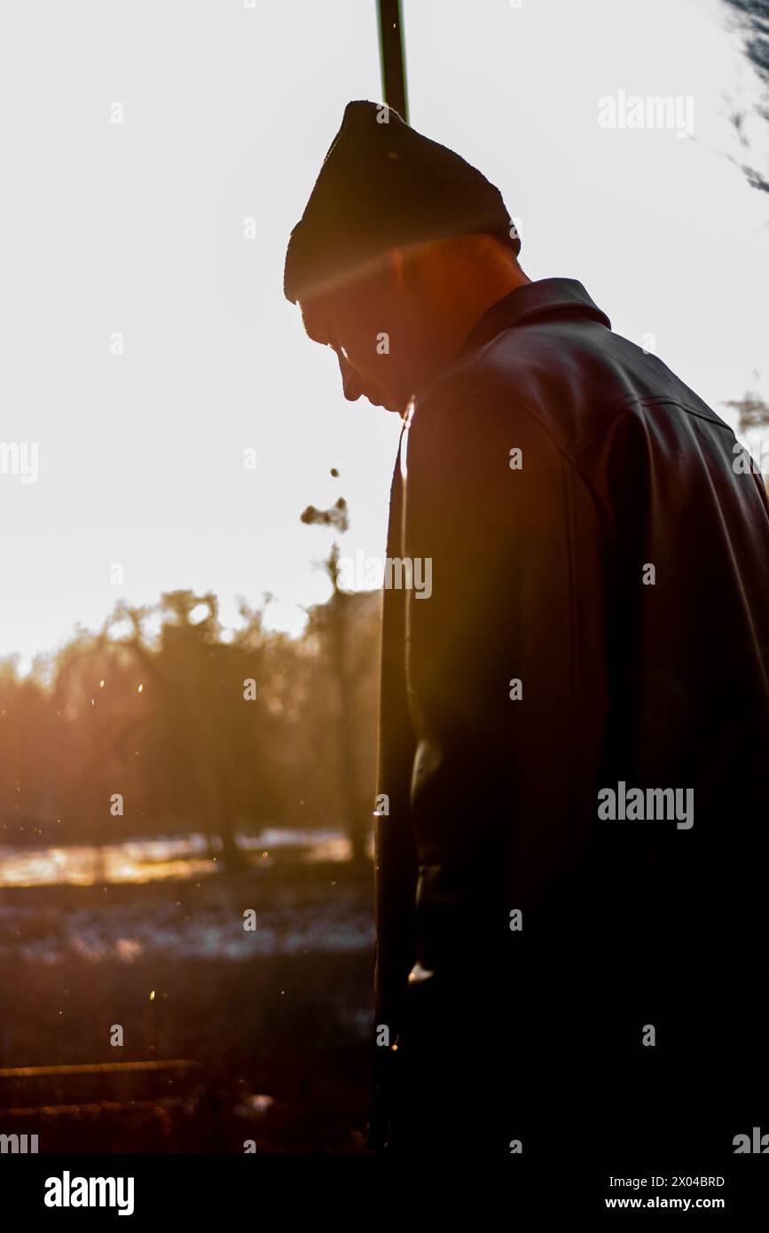 Un bel giovane ragazzo con una giacca e un cappello di pelle nera si posa lateralmente con la testa rivolta verso il basso sullo sfondo dei raggi del sole. Concetto urbano. Foto Stock