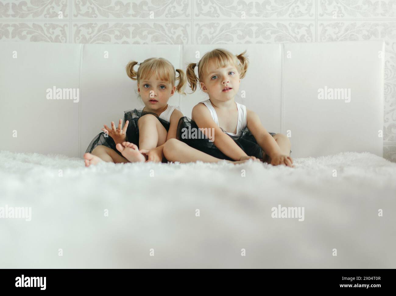 Due ragazze sono sedute fianco a fianco su un letto bianco, guardando qualcosa fuori dalla telecamera. Indossano un abbigliamento casual e sembrano avvolti da wha Foto Stock