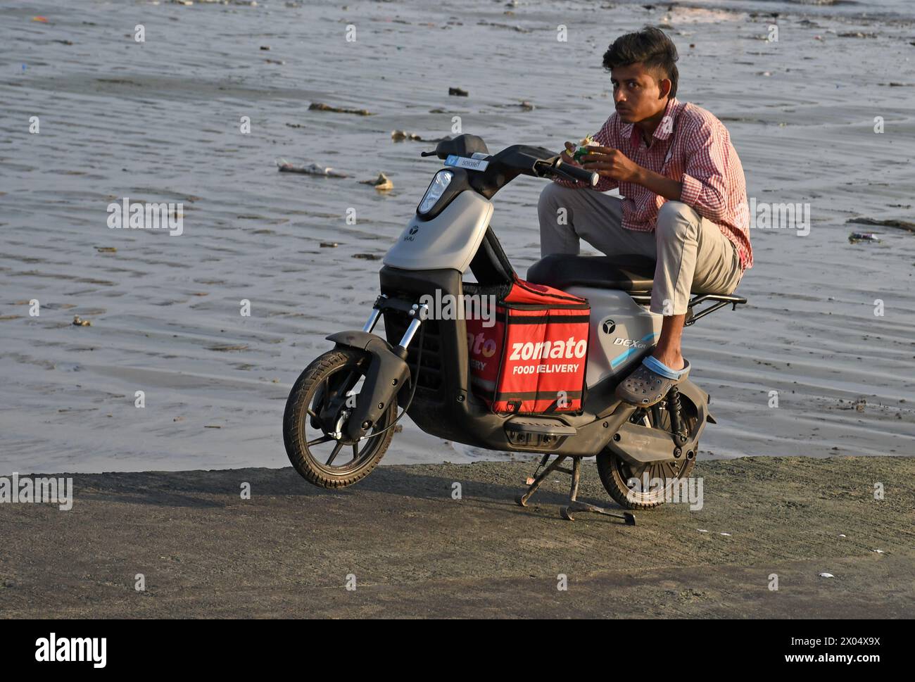 Un addetto alla consegna di Zomato mangia uno spuntino seduto sulla sua bici elettrica in una spiaggia di Mumbai. Zomato è un'azienda di aggregazione di prodotti alimentari che fornisce un'ampia gamma di servizi ai propri clienti online in tutto il paese. Foto Stock