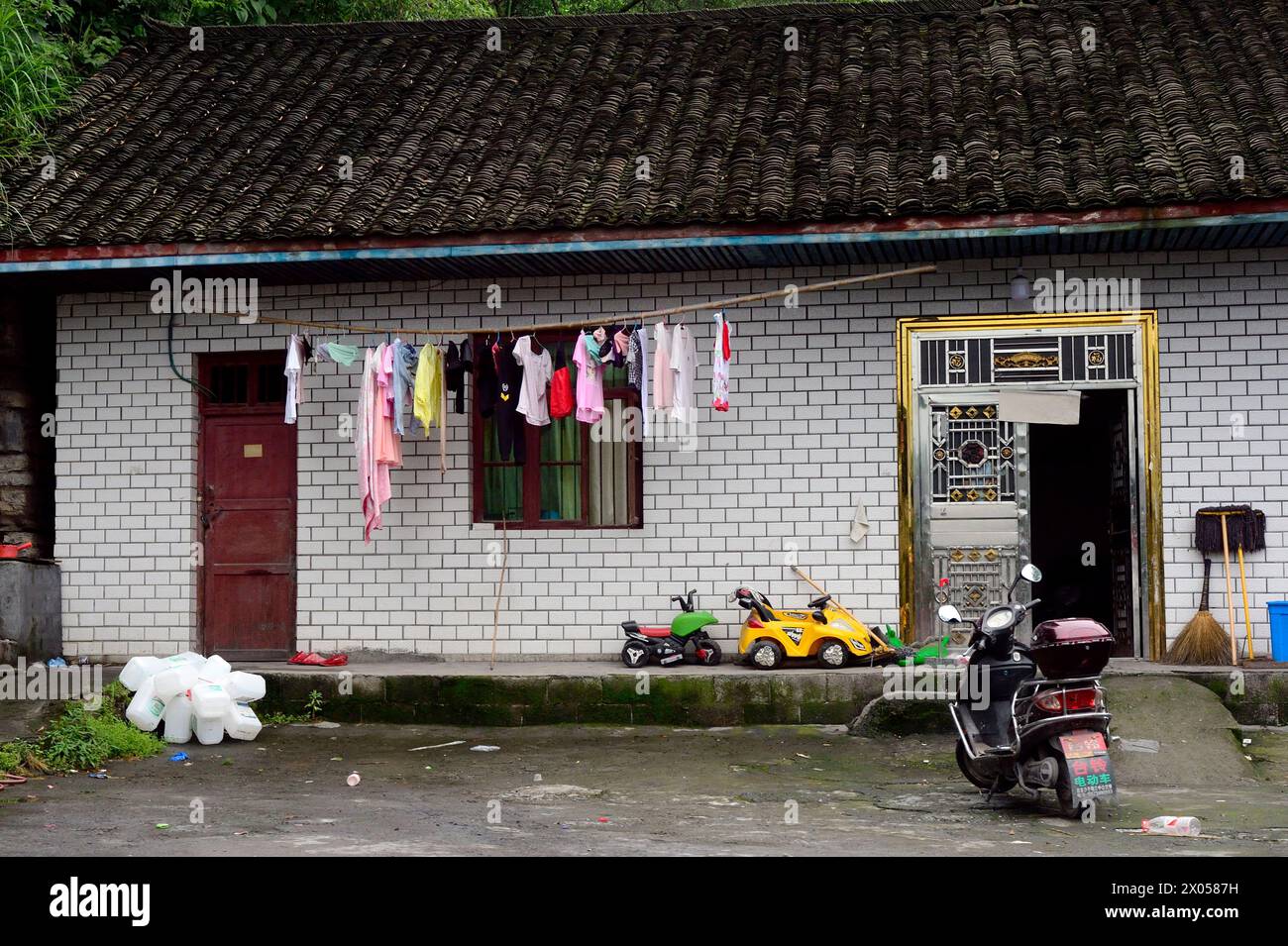 Vestiti per bambini appesi a secco vicino a uno scooter e a una motocicletta giocattolo di fronte a una casa di cemento a Zhangjiajie, in Cina. Foto Stock