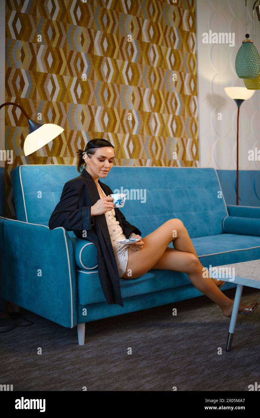 Donna alla moda seduta sul divano con gambe incrociate che guardano con cura in una tazza di tè Foto Stock