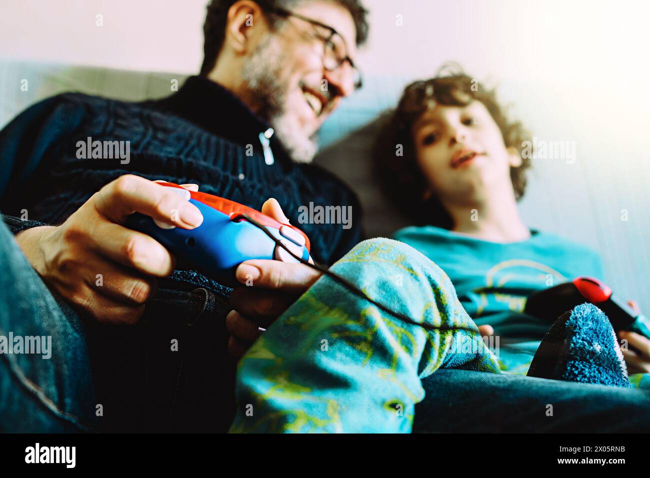 Un padre e un figlio sorridenti si divertono a giocare ai videogame a casa, mentre il padre ha un controller colorato mentre entrambi si concentrano sull'scr Foto Stock