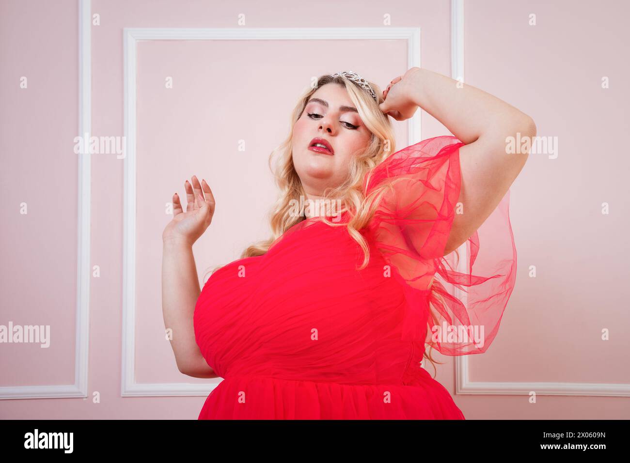 Donna ottimista e curvy in abito rosso con braccio sollevato contro la testa mentre balla dietro sfondo rosa Foto Stock