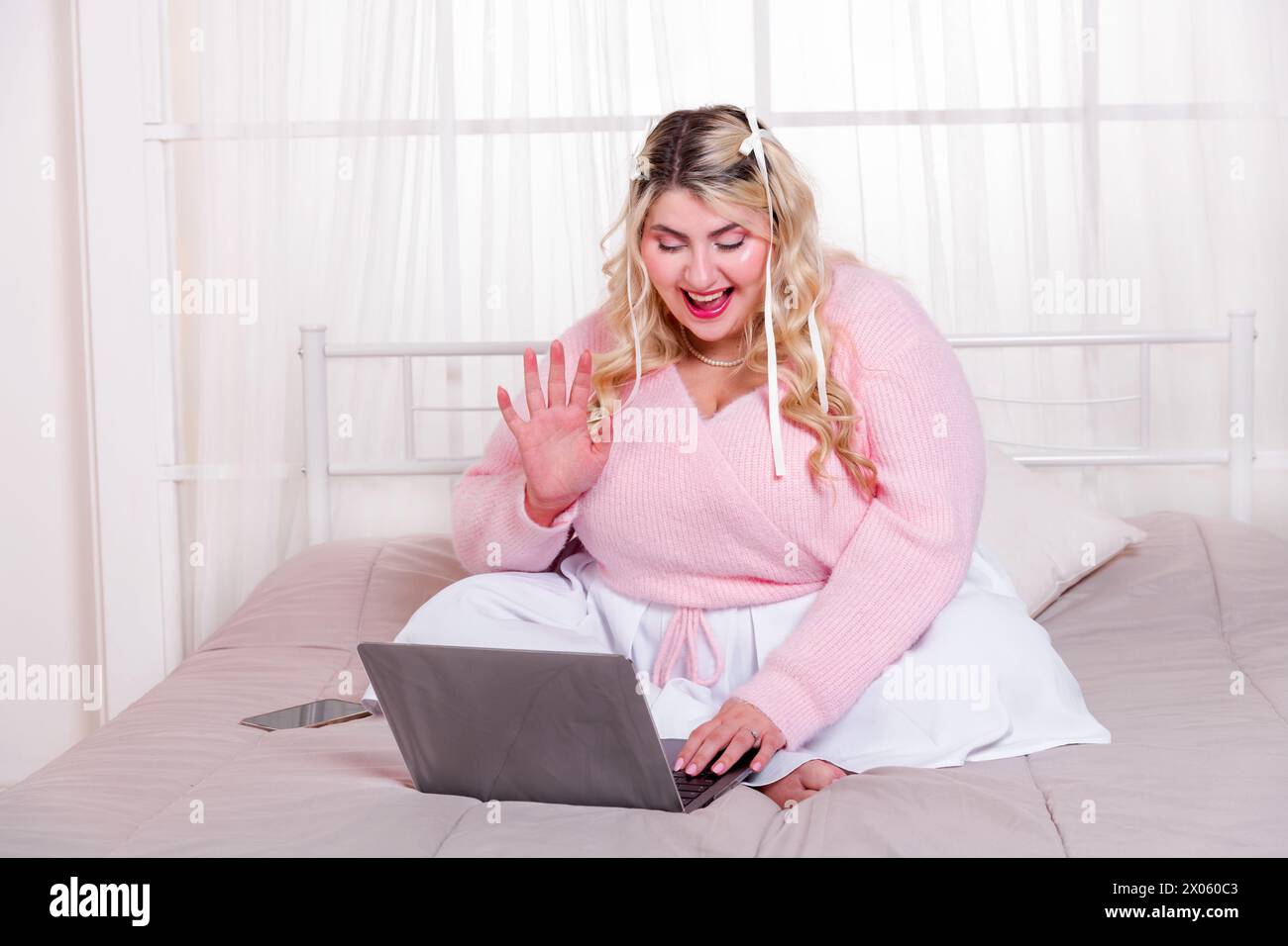 Donna curvy positiva in maglione seduto su un comodo letto che ondeggia la mano mentre utilizza il computer portatile per videochiamare in una stanza accogliente Foto Stock