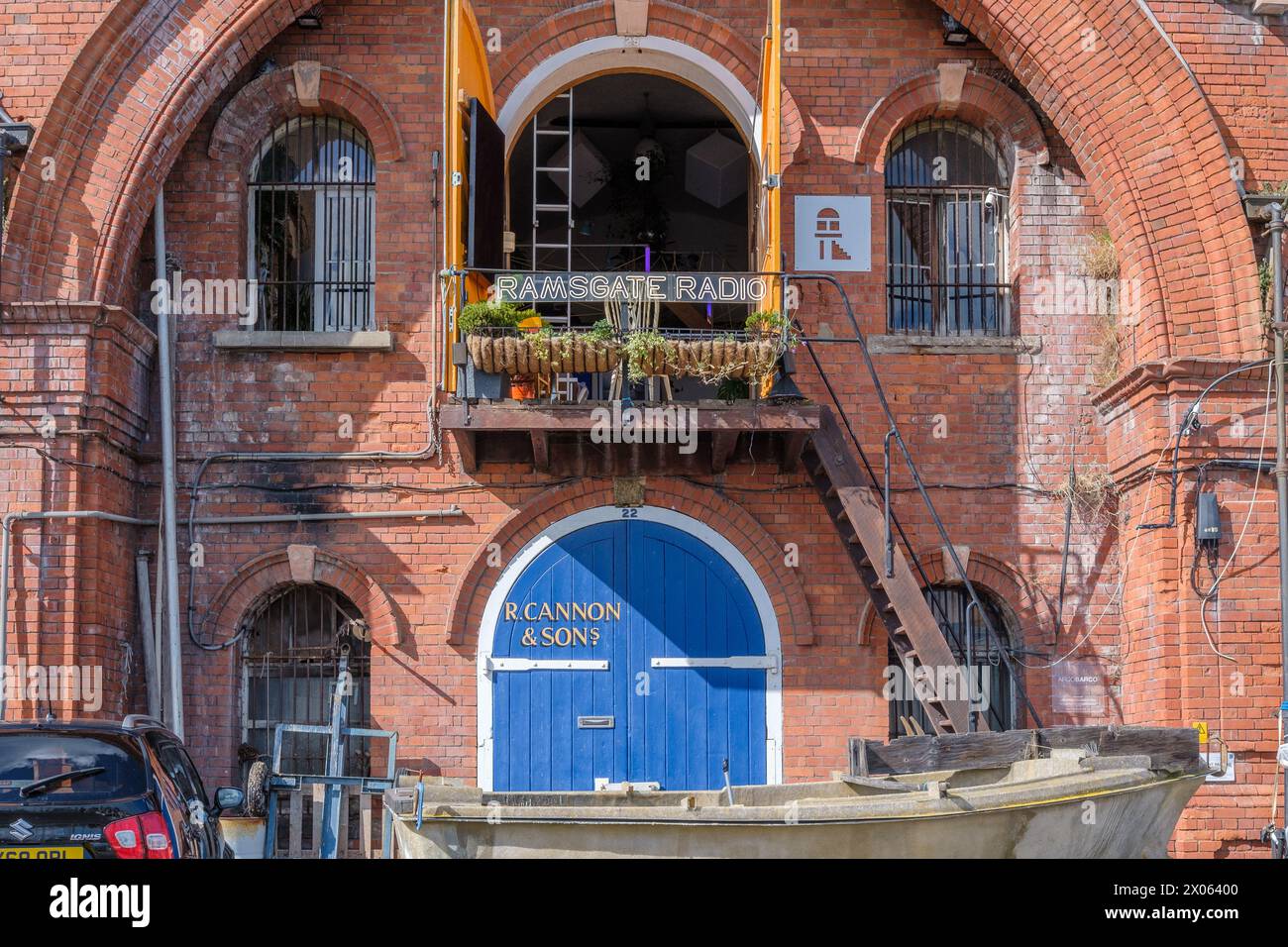 Edificio in mattoni d'epoca con porta blu etichettata R. CANNON & SONS, balcone con bandiere gialle e segnaletica RADIO RAMSGATE. Una vecchia barca a remi di fronte. Foto Stock