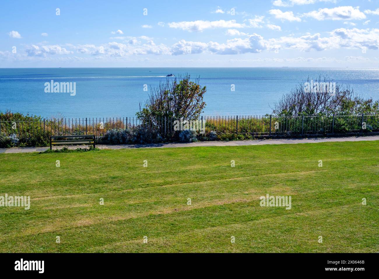 La panca in legno si affaccia sul mare calmo sotto il cielo limpido. Il primo piano è ricoperto di lussureggiante erba verde. La recinzione confina con l'area. Thanet Coast Kent, Regno Unito. Foto Stock