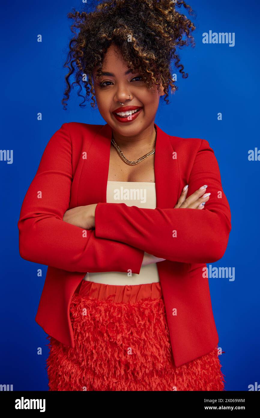 allegra e seducente donna afroamericana in rosso blazer con capelli raccolti sorridenti alla macchina fotografica Foto Stock