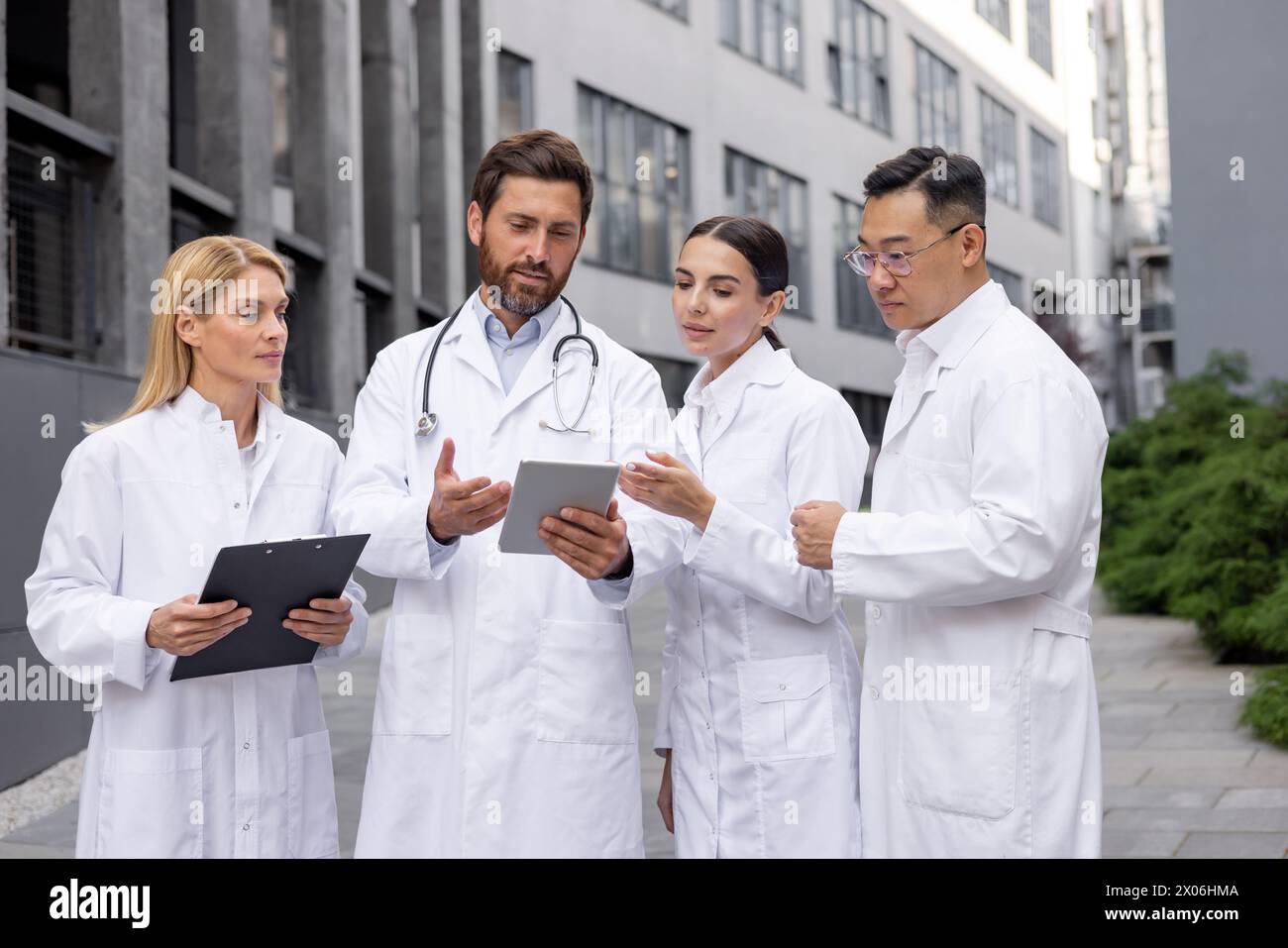 Un gruppo di giovani medici e scienziati interrazziali maschili e femminili si trova fuori da un ospedale in cappotti bianchi e discutere dati e ricerche su una compressa. Foto Stock