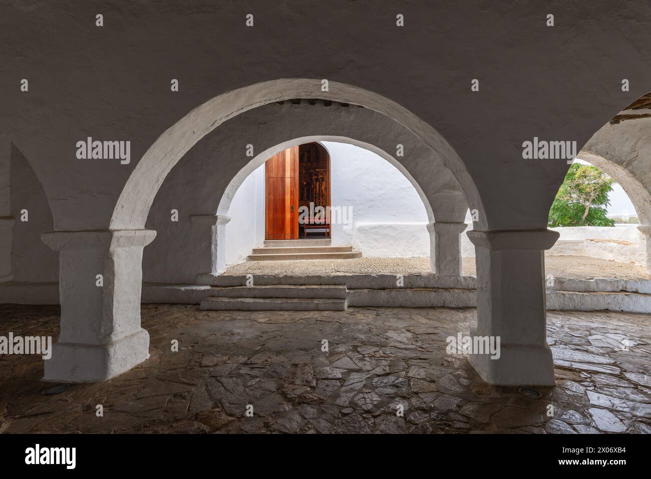 L'atmosfera serena del cortile coperto della chiesa di Puig de Missa a Santa Eulalia, Ibiza, con i suoi archi bianchi che incorniciano una vista sulla porta di legno Foto Stock