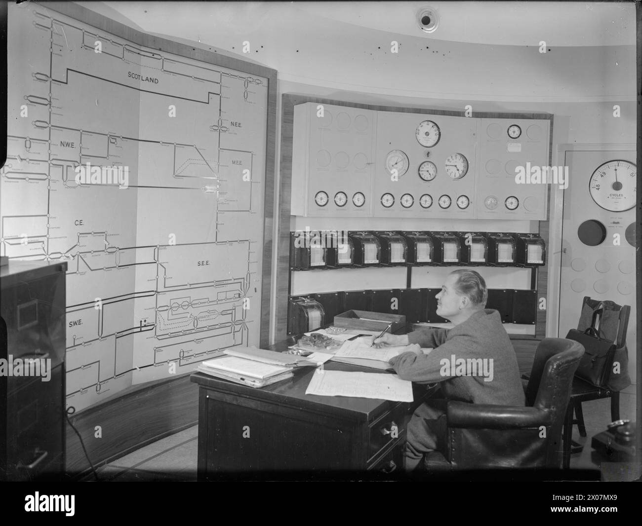 IL SISTEMA DI RETE INGLESE: IL LAVORO DEL CONSIGLIO CENTRALE DELL'ELETTRICITÀ IN TEMPO DI GUERRA, c MARZO 1945 - Un controllore al lavoro nella sala centrale di controllo dell'elettricità. Sulla sinistra è presente uno schema del sistema che mostra l'impianto elettrico per l'intera Gran Bretagna, dalla Scozia in alto, passando per "NWE" (Inghilterra nord-occidentale), "CE" (Inghilterra centrale) e "SWE" (Inghilterra sud-occidentale), tra gli altri. Secondo la didascalia originale, questa sala di controllo è ben al di sotto del suolo, quindi è al sicuro da qualsiasi attacco nemico ed è da qui che "l'alimentazione può essere commutata da aree meno cariche a industrie di guerra vitali" Foto Stock