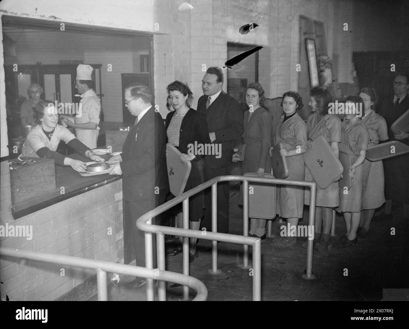 BILLY BUTLIN VISITA Una FABBRICA DI MUNIZIONI, Regno Unito, 1941 - il signor Butlin raccoglie un pasto dal portello di servizio nella mensa di un ostello di una fabbrica di munizioni, da qualche parte in Gran Bretagna. Le lavoratrici fanno la fila dietro di lui per ritirare il loro pranzo Butlin, Wiliam Heygate Edward Colborne Foto Stock