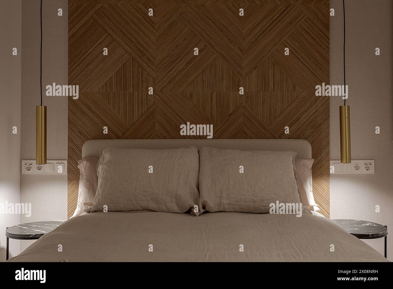 Una camera da letto accogliente ed elegante con enfasi su una suggestiva testata in legno caratterizzata da un motivo a spina di pesce, fiancheggiata da moderne luci a incandescenza Foto Stock