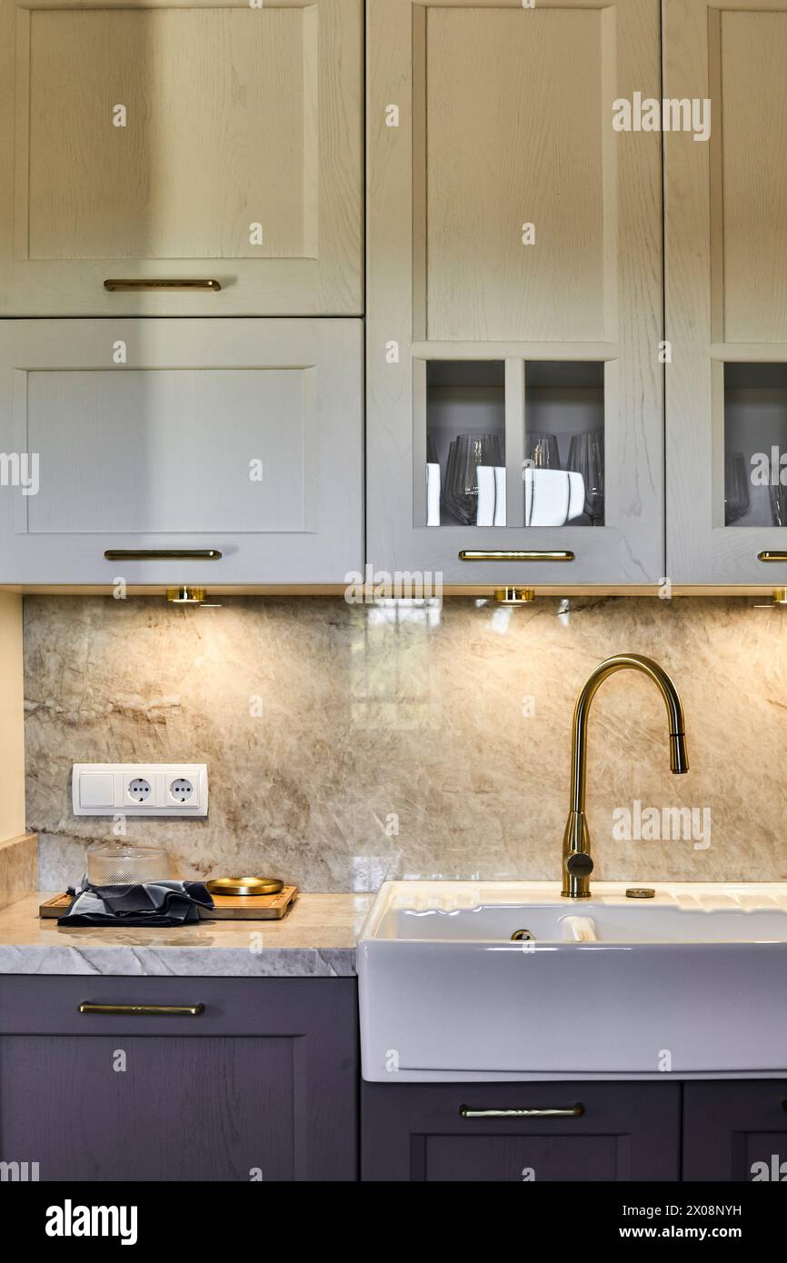Cucina dal design moderno ed elegante con lavabo bianco della fattoria con rubinetto in ottone, adagiato su uno splendido lavabo in marmo e armadietto in legno chiaro Foto Stock