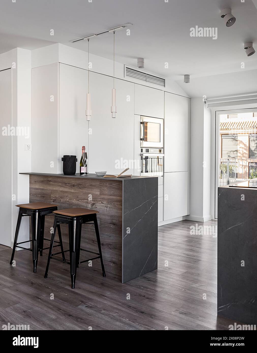 Un'elegante cucina moderna con mobili bianchi, accenti in legno e sgabelli neri, completati da luce naturale Foto Stock