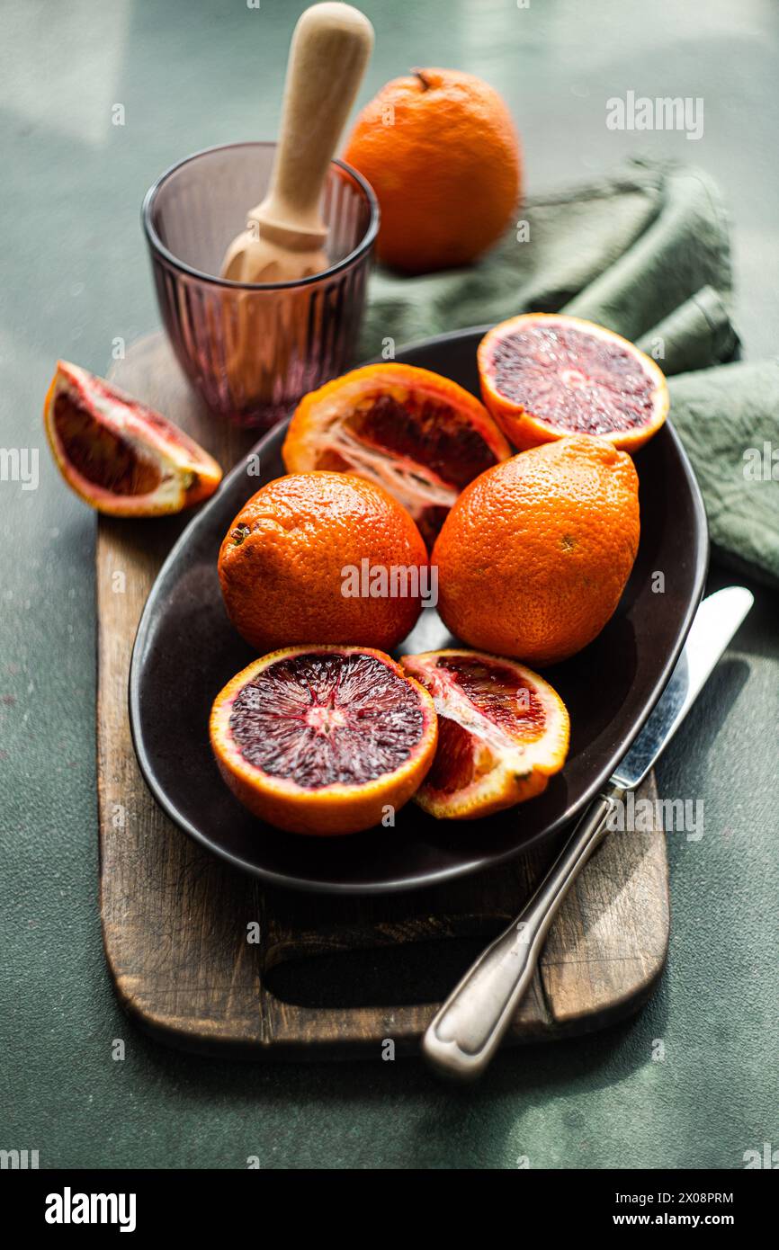 Le succose arance di sangue si sono dimezzate e poste su un piatto scuro, con un tovagliolo verde e una centrifuga di agrumi sullo sfondo Foto Stock