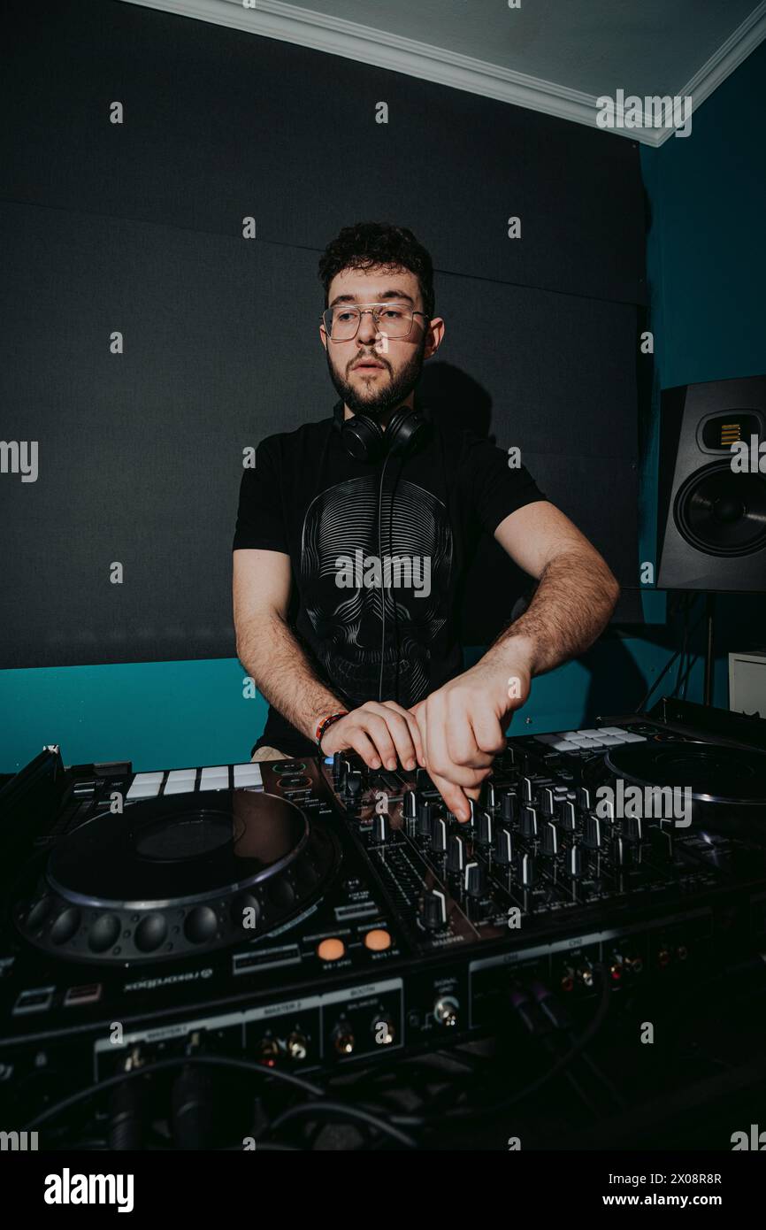 Un giovane DJ maschile si concentra mentre mescola i brani su un mixer audio professionale in un ambiente da club, con un'atmosfera scura e luci colorate Foto Stock
