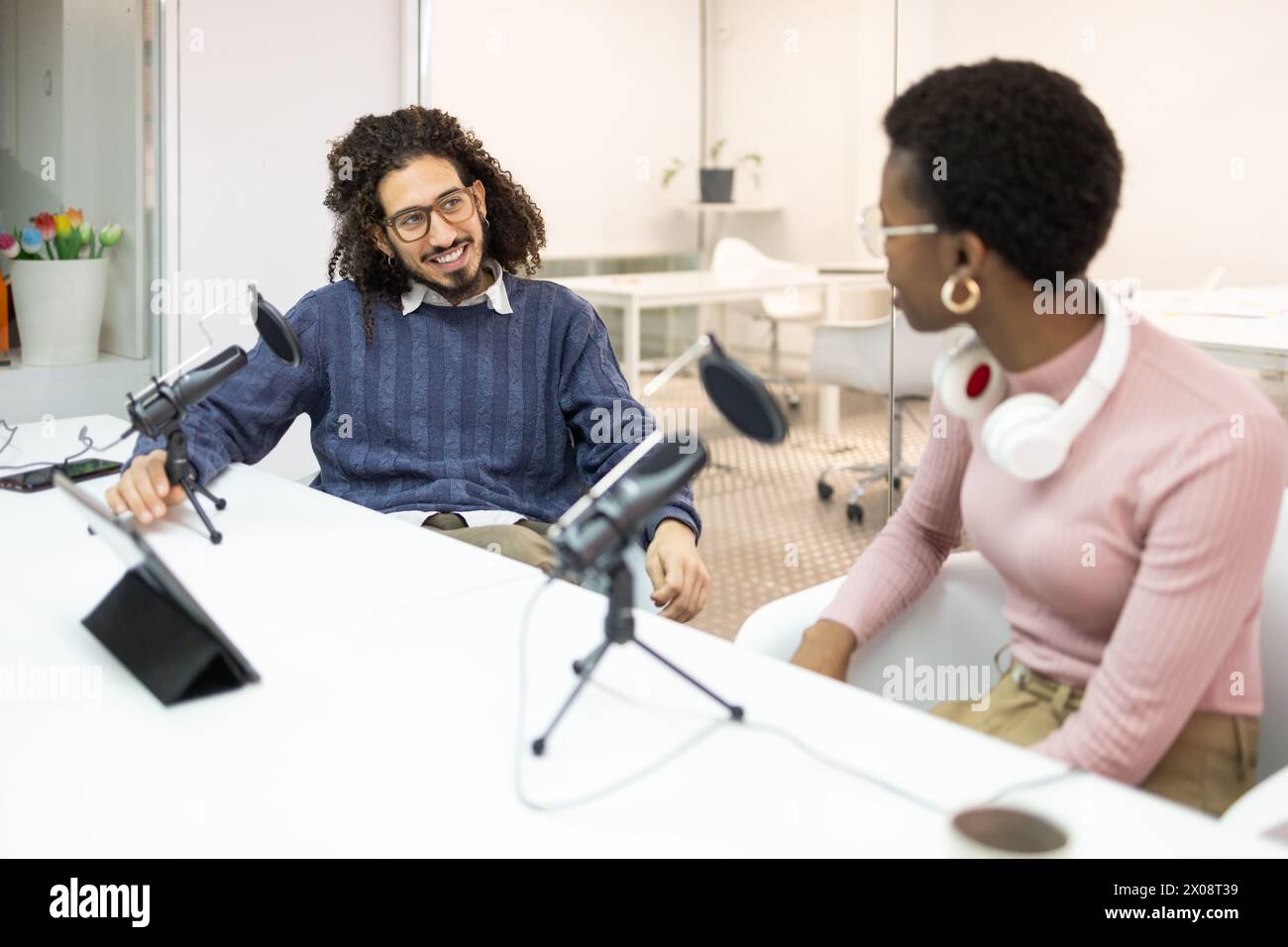 Un team di podcast multirazziale durante una sessione di registrazione in un ambiente luminoso in studio. La donna, forse afroamericana, sta guardando il suo co- Foto Stock