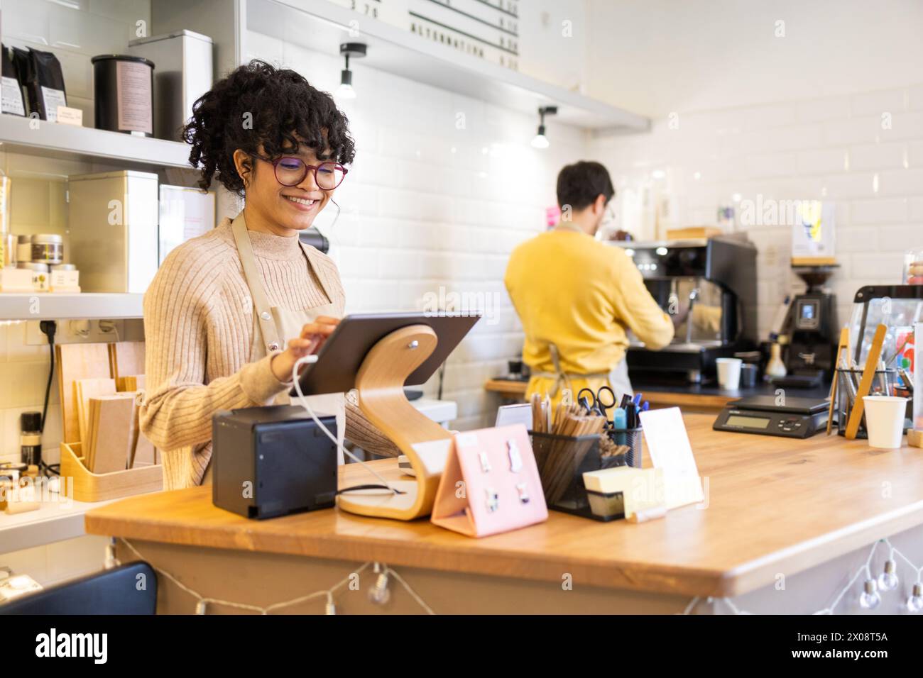La barista sorridente ispanica gestisce un tablet digitale presso un moderno bancone di caffetteria, con un collega che prepara bevande in sottofondo. Foto Stock