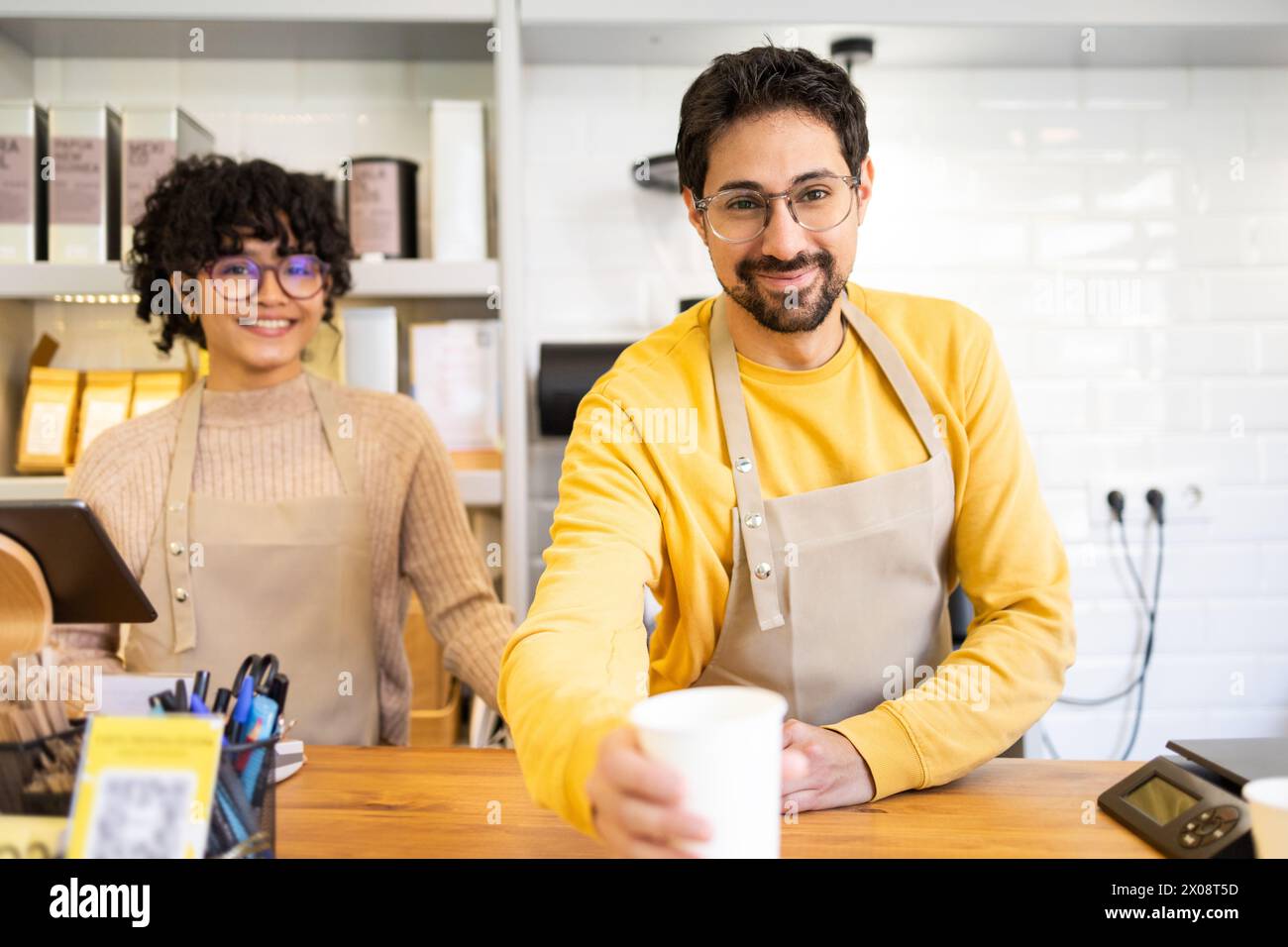 Due allegri baristi multietnici con sorrisi caldi in piedi dietro il bancone in un caffè moderno, che offre una tazza di caffè appena fatto. Foto Stock