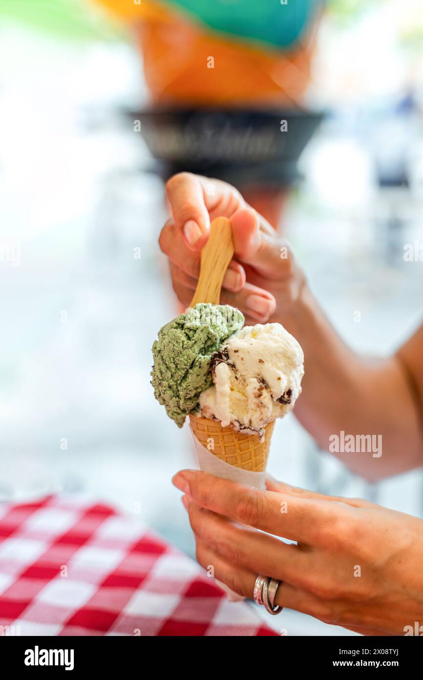 Uno scatto ravvicinato che cattura la mano di una persona irriconoscibile ritagliata che tiene in mano un cono con due palline di gelato, contro un retrogroun vibrante e sfocato Foto Stock