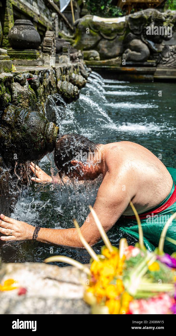 Una persona si impegna in una cerimonia di purificazione dell'acqua in una sorgente sacra, ambientata contro statue intricate scolpite, a Bali. Foto Stock