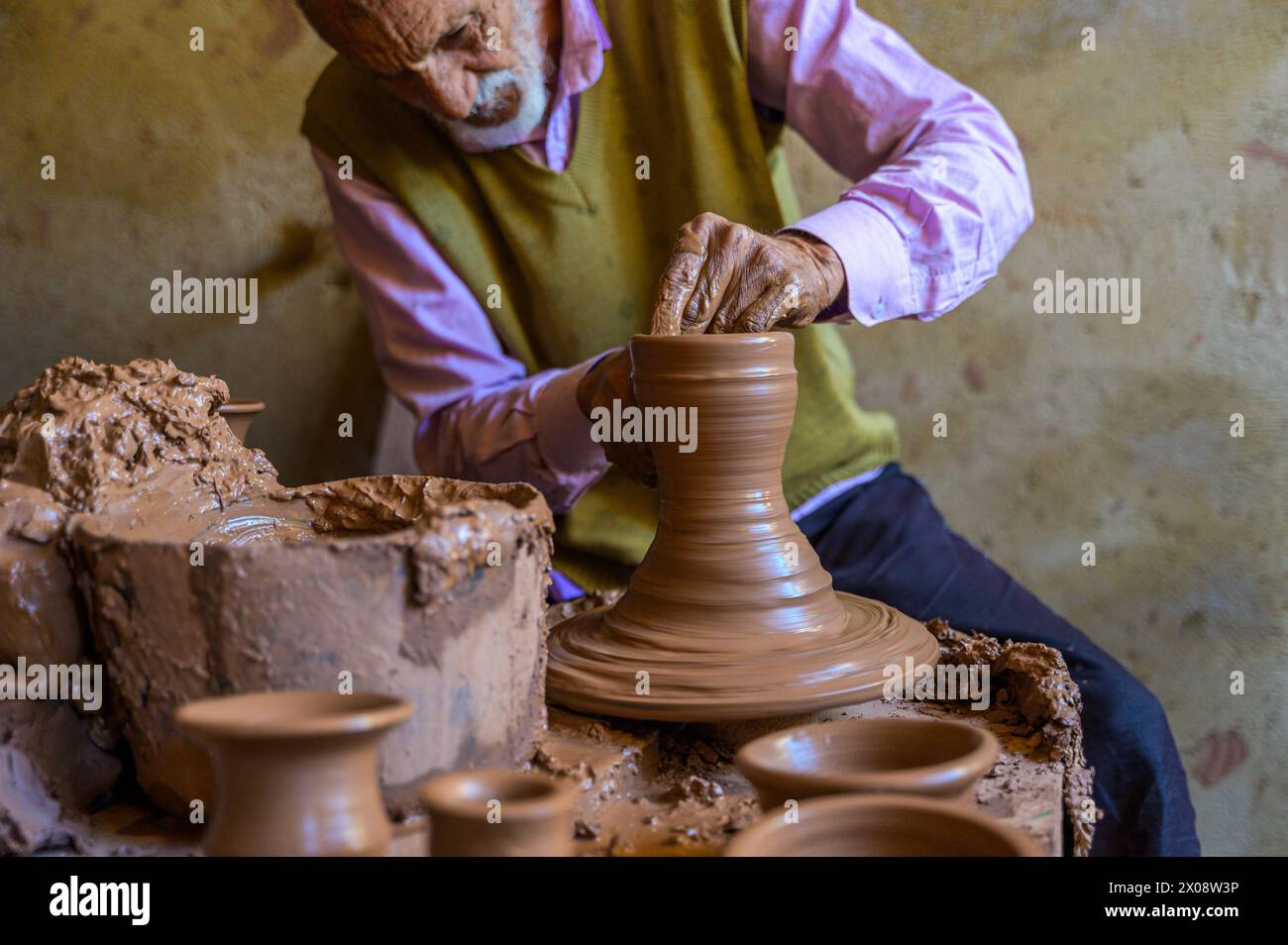 Un anziano vasaio esperto forma un vaso di argilla bagnato su una ruota di ceramica rotante in un laboratorio rustico, che esemplifica l'artigianato artigianale. Foto Stock