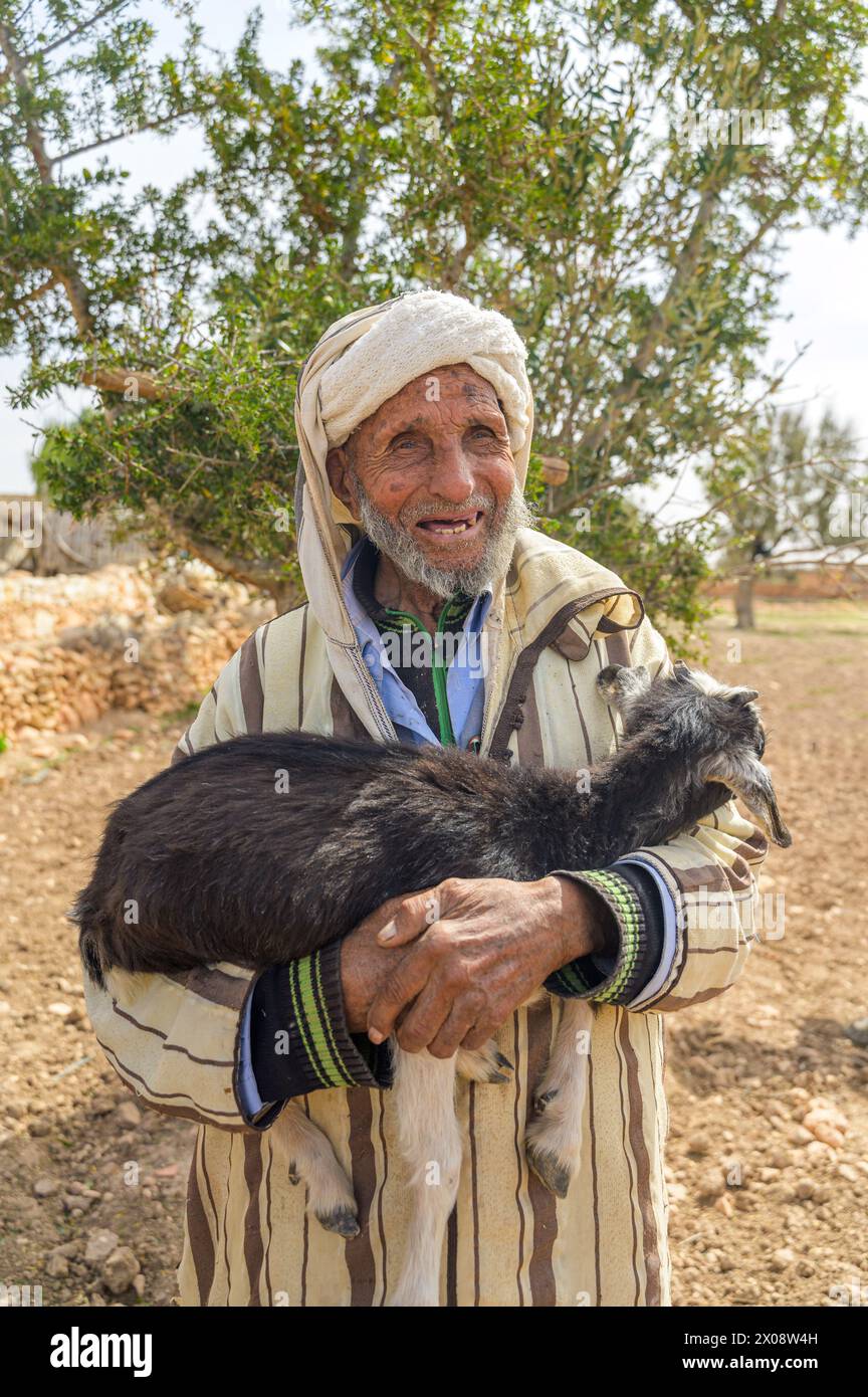 Un anziano marocchino in abbigliamento tradizionale sorridente mentre culla una capra nera in un paesaggio rurale, mostrando lo stile di vita e la cultura locali Foto Stock