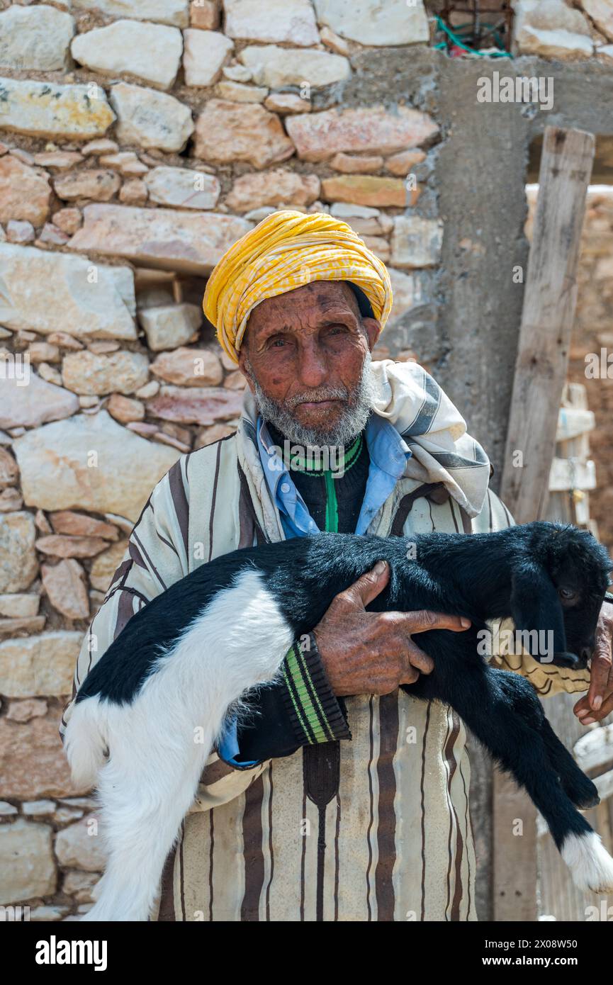 Un anziano marocchino, con indosso un tradizionale turbante giallo, tiene affettuosamente una giovane capra bianca e nera davanti a un muro di pietra Foto Stock