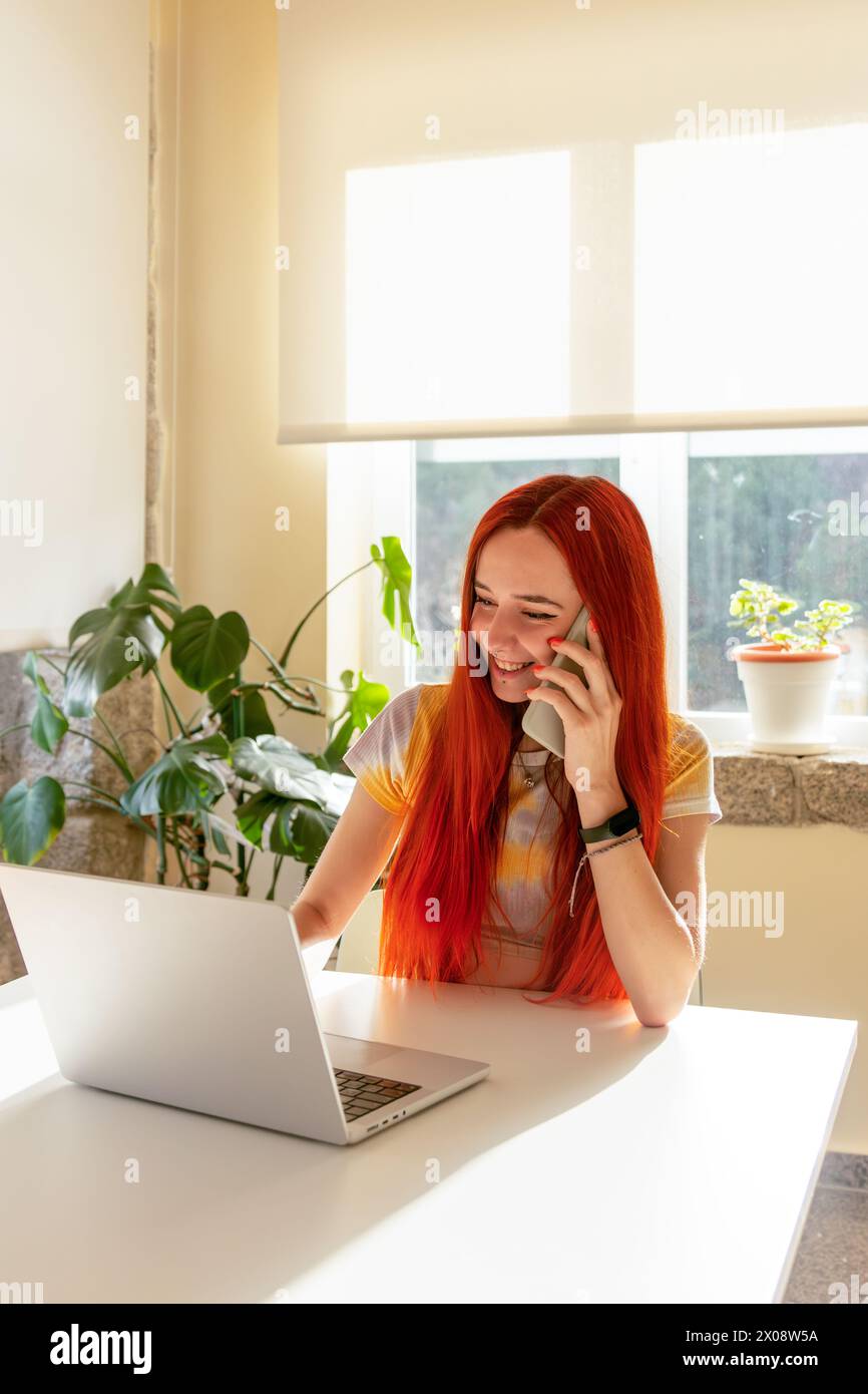 Una donna sorridente dalla testa rossa che sta pianificando una vacanza nella natura al telefono, con un laptop aperto davanti a lei in una stanza luminosa e illuminata dal sole Foto Stock