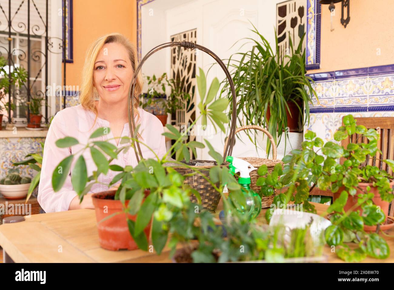 Una donna allegra siede su un tavolo di legno circondato da lussureggianti piante in vaso su un patio luminoso e decorato. Foto Stock