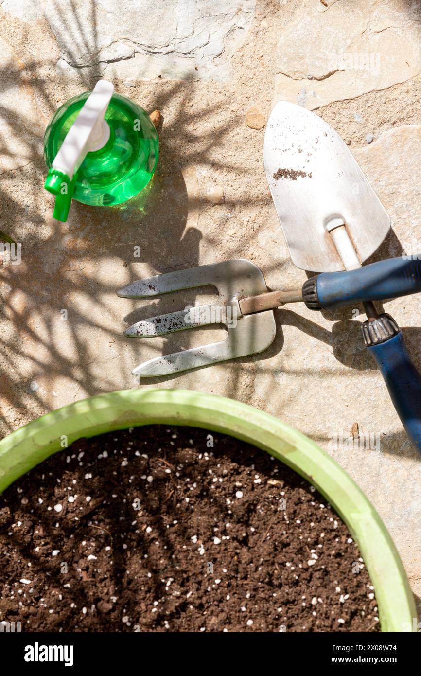 Attrezzi da giardinaggio disposti su un patio, tra cui una lattina da irrigazione verde, una vanga e una forchetta manuale, con una pentola riempita di terreno pronta per la semina. Foto Stock
