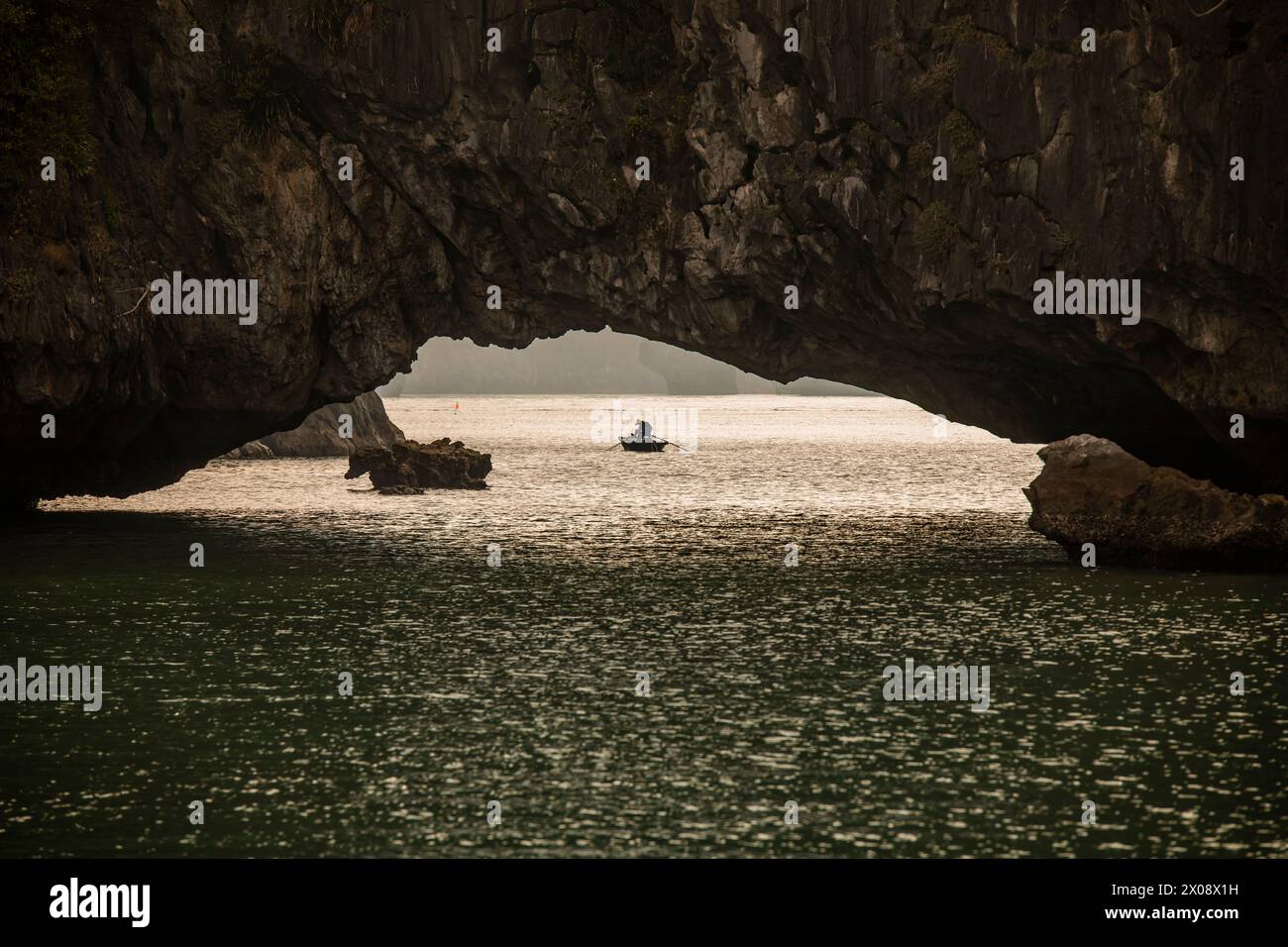 Una barca tradizionale con pescatori irriconoscibili scivola attraverso le tranquille acque della baia di ha Long, in Vietnam, passando sotto un arco calcareo naturale con Foto Stock