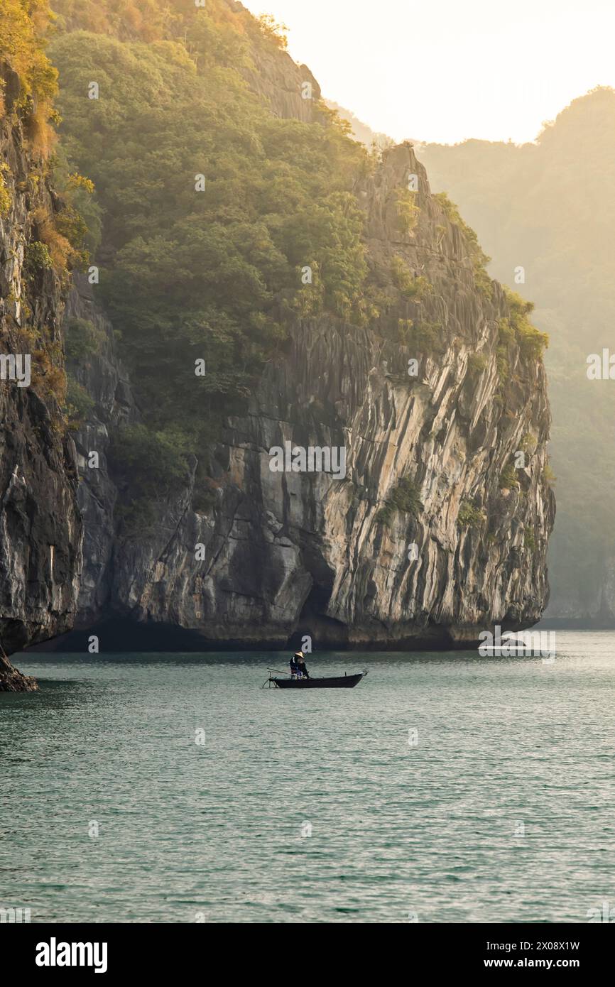 Una barca solitaria con pescatori irriconoscibili naviga nelle tranquille acque della baia di ha Long, incorniciata da torreggianti scogliere calcaree bagnate dalla l dorata Foto Stock