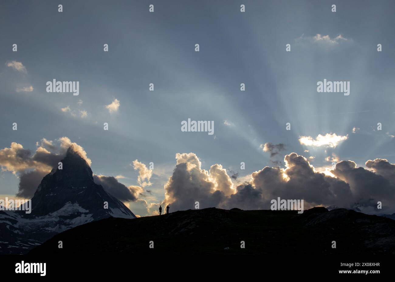 Due escursionisti si stagliano contro un cielo dove le onde del sole si infrangono attraverso le nuvole sopra il Cervino al crepuscolo, mettendo in risalto la bellezza della Swiss al Foto Stock