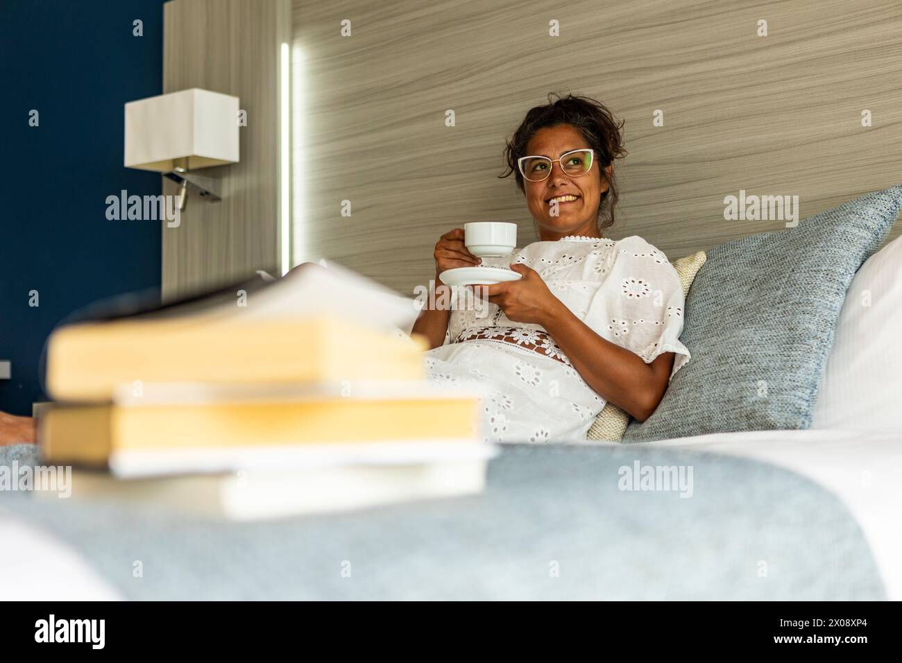 Una donna ispanica sorridente di mezza età con i bicchieri giace a letto con una tazza di caffè, con una pila di libri in primo piano. Foto Stock