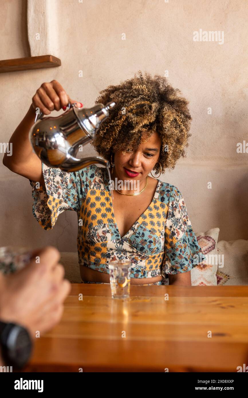 Una donna allegra che versa l'acqua da una caraffa in un bicchiere per accompagnarla in un bar caldo e invitante, simboleggiando l'amicizia e il tempo libero Foto Stock