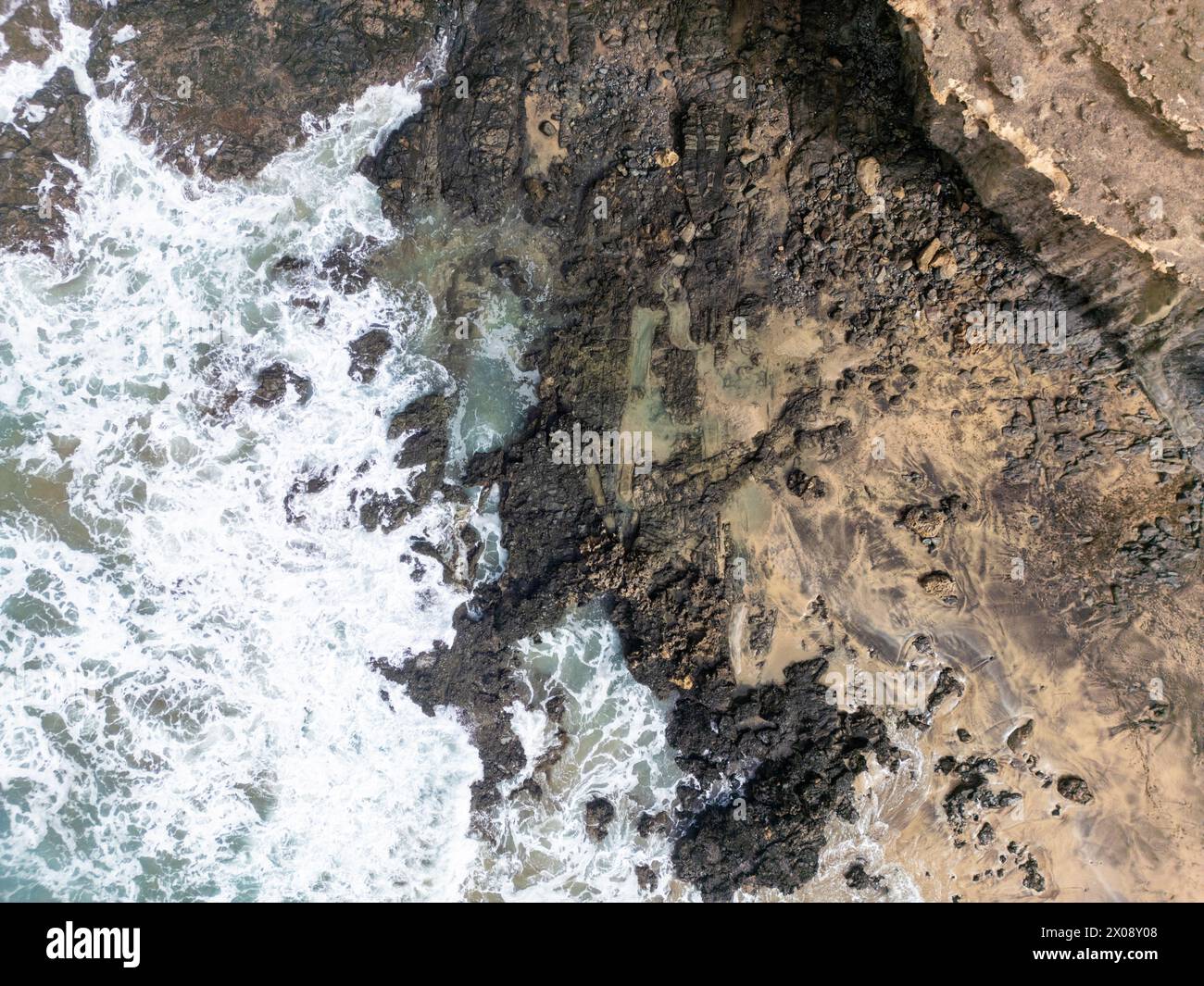 Scatto aereo che cattura l'interazione dinamica tra le onde bianche e spumeggianti e la costa rocciosa a Cofete Beach, Costa calma a Fuerteventura Foto Stock