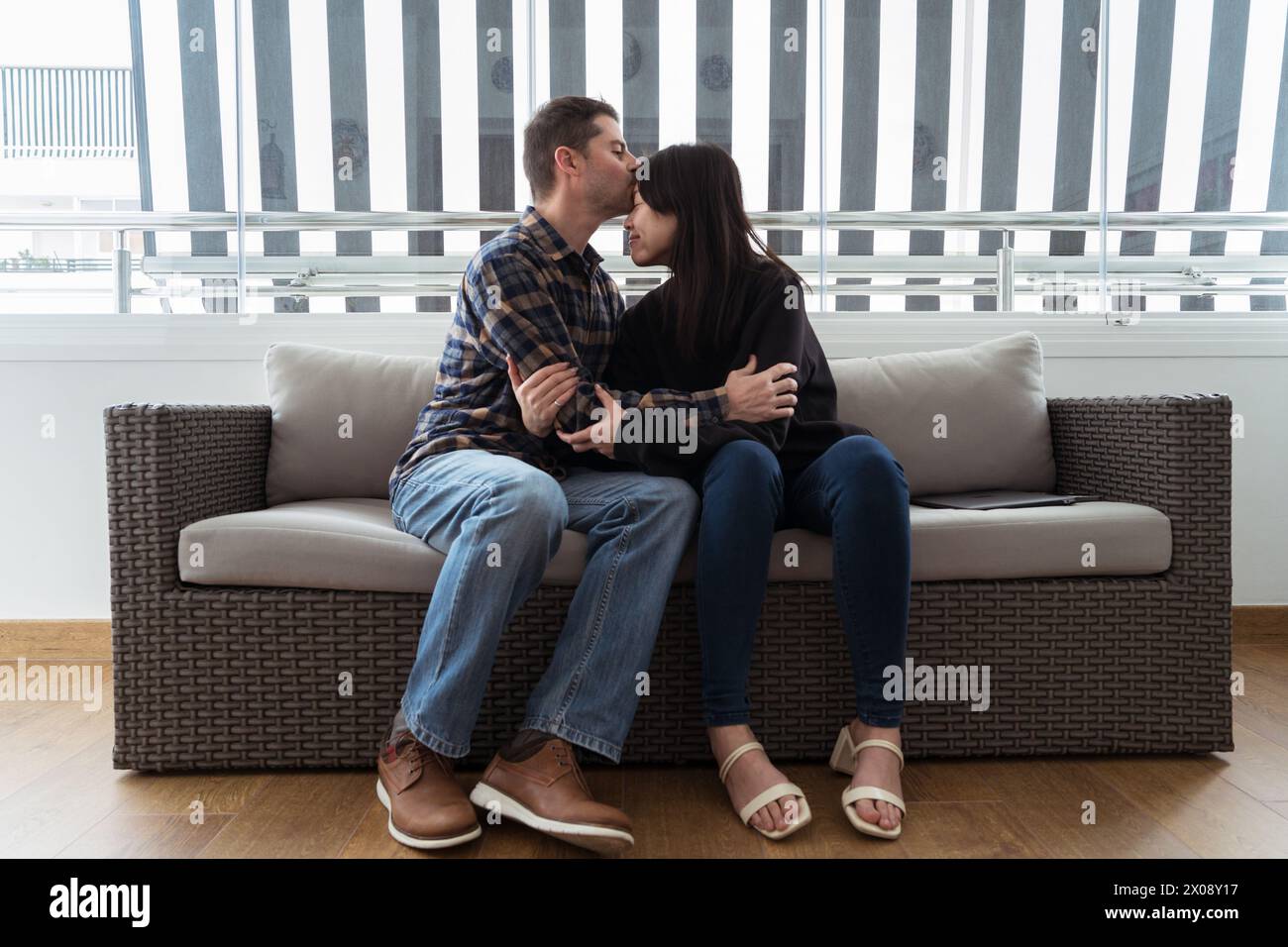 Una donna asiatica e un uomo caucasico abbracciano e baciano la fronte su un divano che mostra amore e compagno in tutte le culture Foto Stock