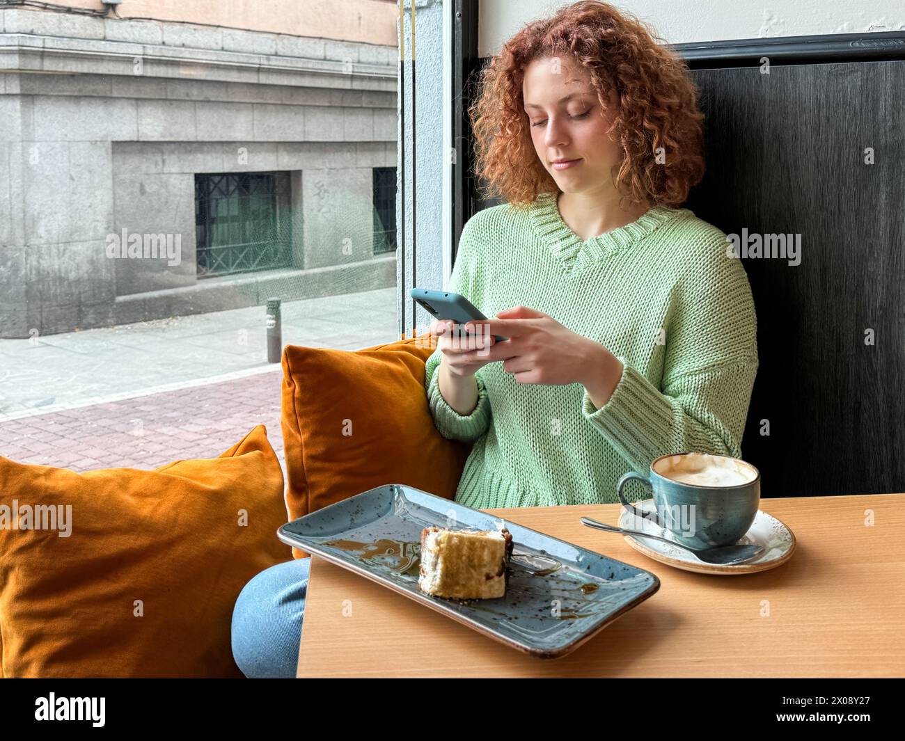 Giovane donna rossa dai capelli ricci seduta a un tavolino vicino a una finestra, usando uno smartphone con una tazza di caffè e una fetta di torta Foto Stock