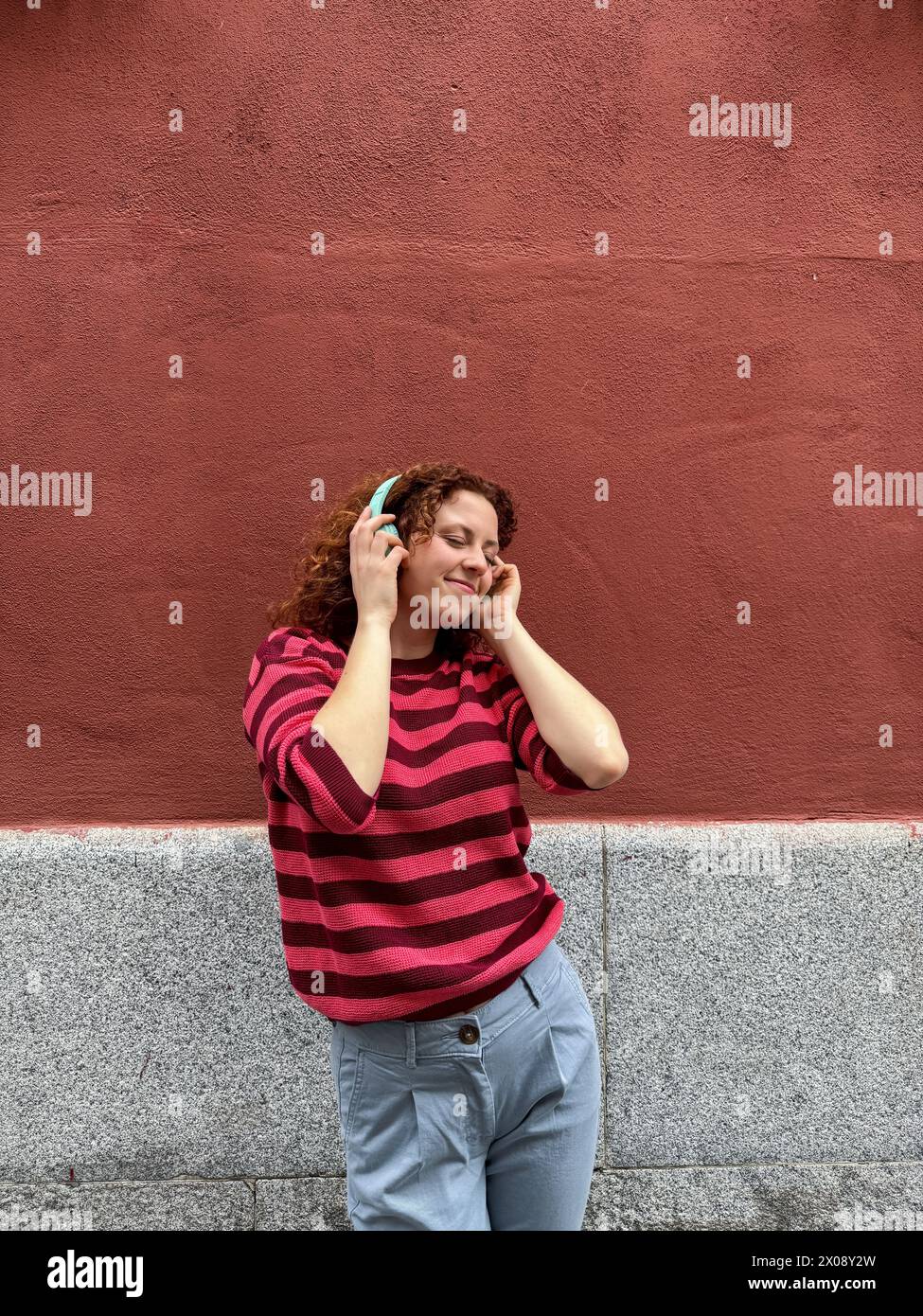 Una giovane donna rossa allegra con i capelli ricci sorride mentre ascolta la musica con le sue cuffie color acqua contro un muro rosso Foto Stock