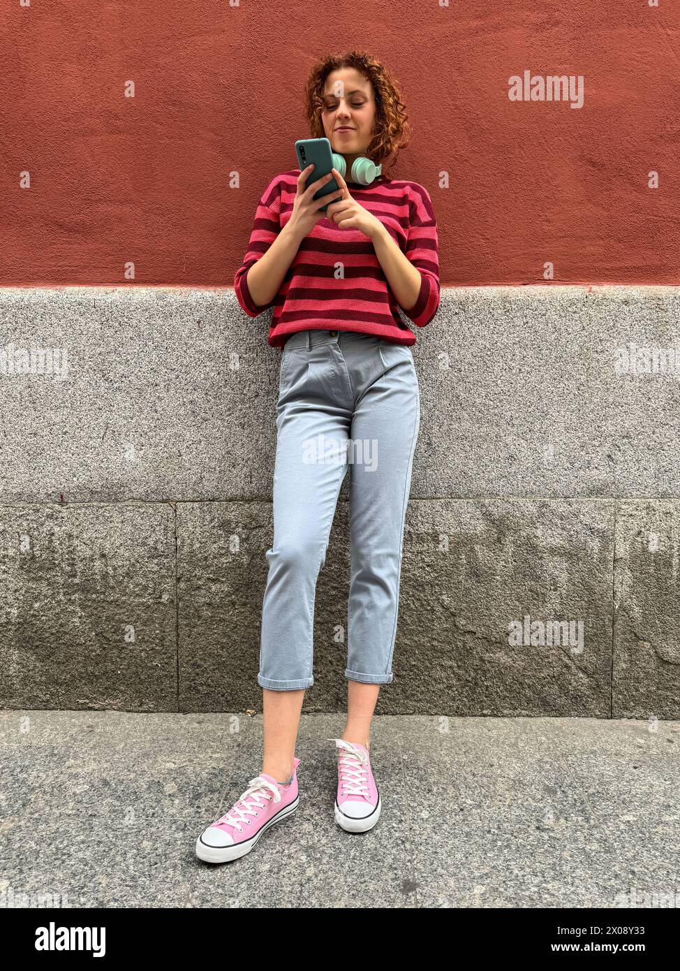 Una giovane donna rossa dai capelli ricci in piedi e che sfoglia il telefono con le cuffie intorno al collo contro una vivace parete rossa Foto Stock