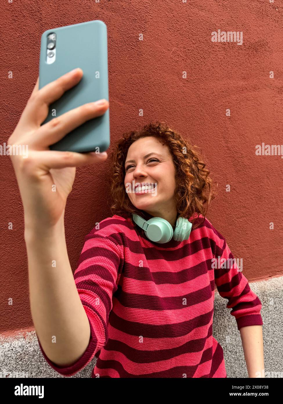 Una donna rossa allegra con i capelli ricci sorride mentre scatta un selfie, indossando una camicia a righe e cuffie intorno al collo Foto Stock
