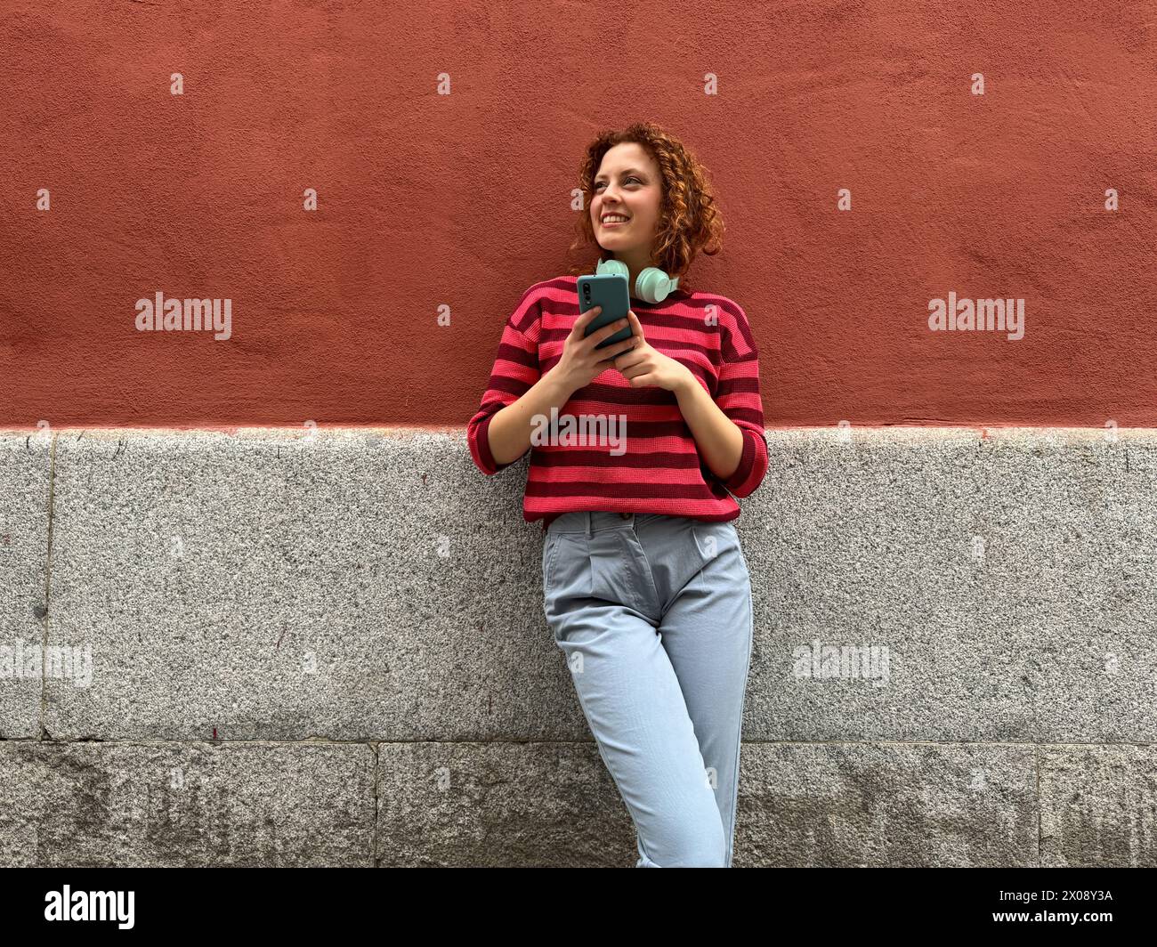 Una donna rossa sorridente con i capelli ricci si appoggia contro un muro, si impegna con il telefono e con le cuffie intorno al collo in una giornata di sole Foto Stock