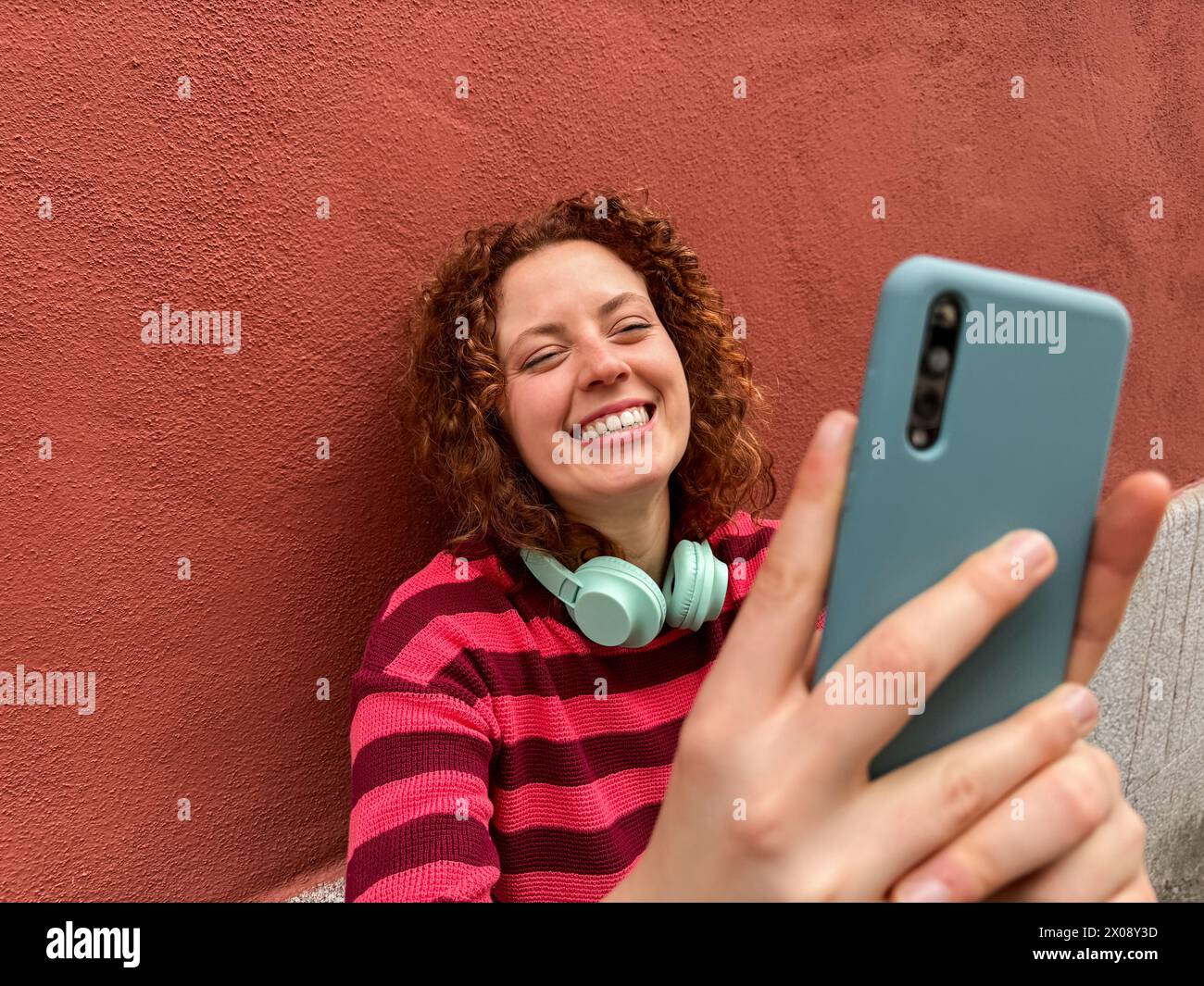 Sorridente giovane rossa con maglione a righe che fa una divertente video chat sul suo smartphone su uno sfondo rosso Foto Stock
