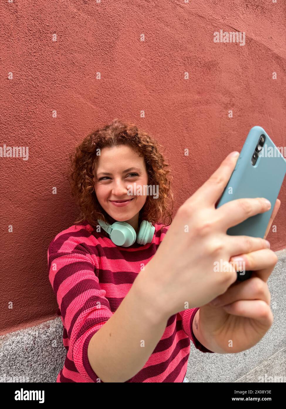 Un'allegra donna rossa dai capelli ricci con una camicia a righe scatta un selfie con il suo smartphone, indossando le cuffie intorno al collo Foto Stock