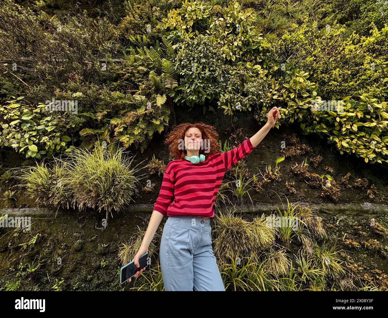 Una giovane donna rossa gioiosa con capelli ricci si allunga le braccia in mezzo a una vivace parete verde di piante, incarnando libertà e felicità Foto Stock
