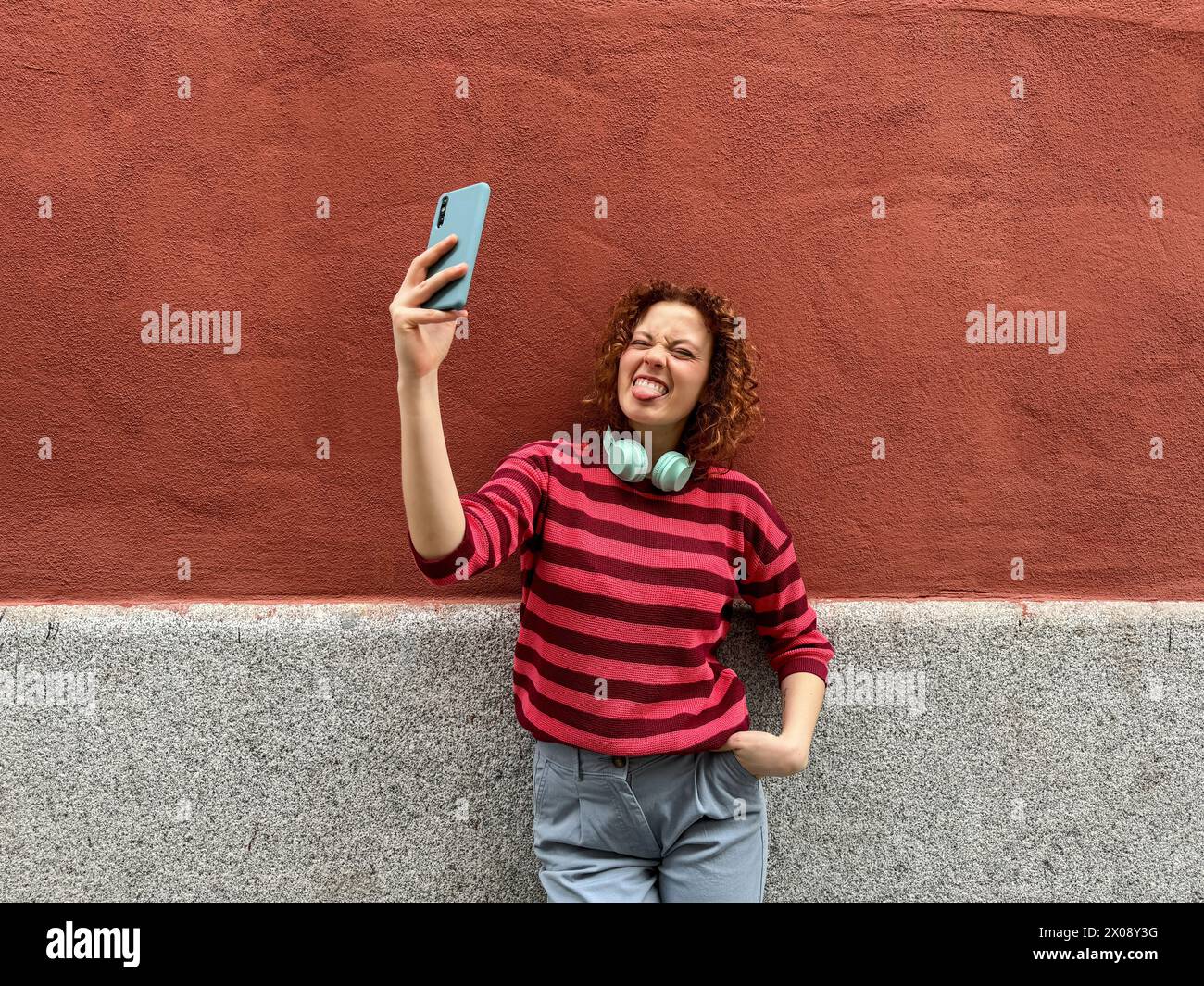 La giovane donna allegra con una camicia a righe e le cuffie si attacca scherzosamente con la lingua mentre scatta un selfie contro un muro rosso Foto Stock