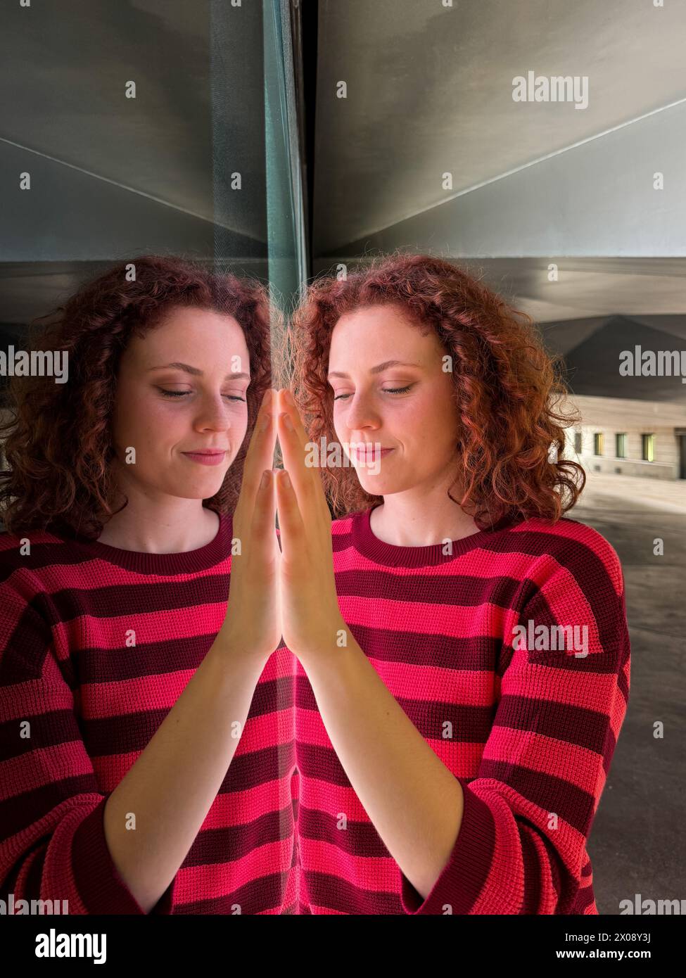 Una donna rossa con una camicia a righe rosse che si riflette su una superficie specchiata, creando un'immagine simmetrica Foto Stock