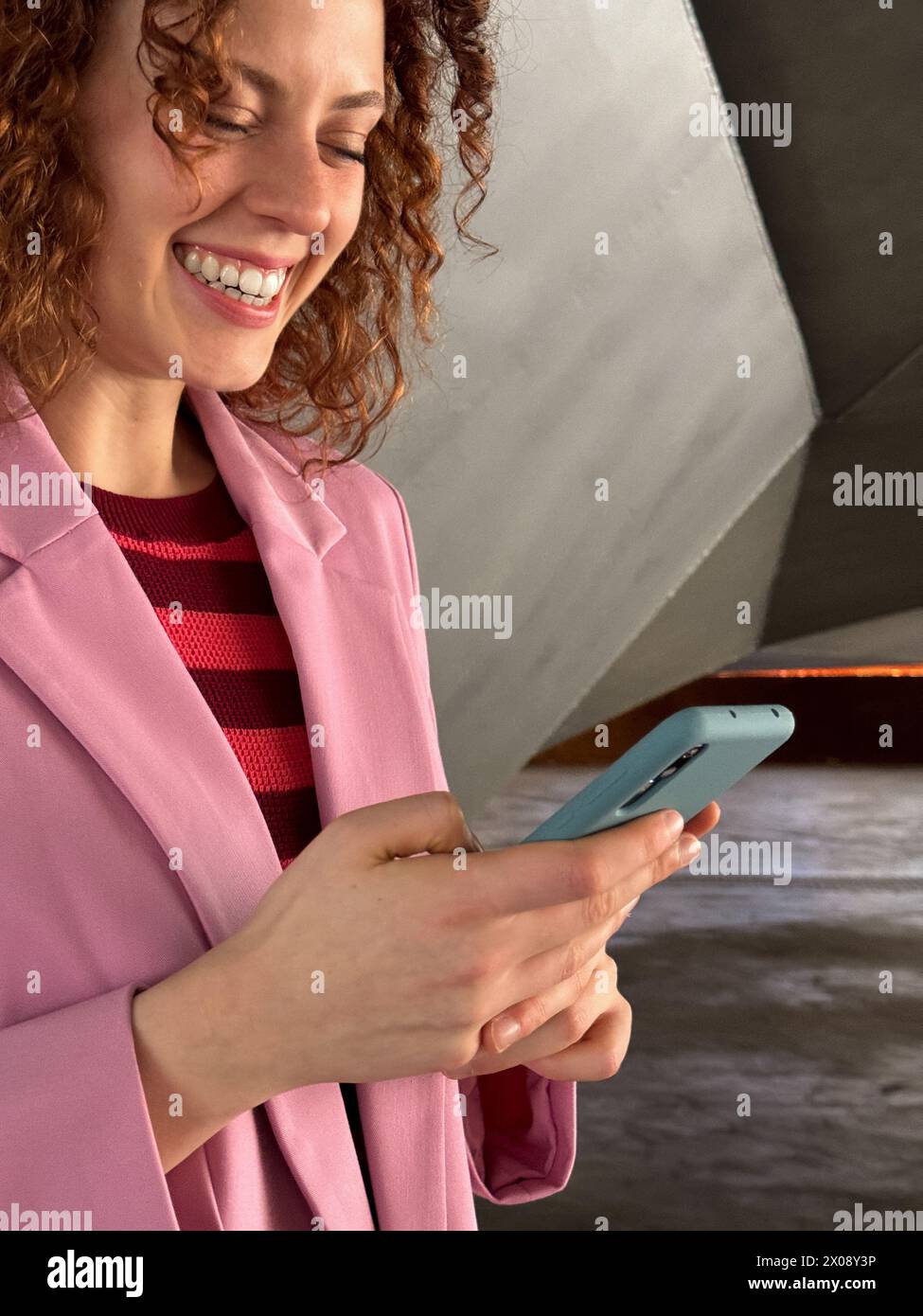 Primo piano di una gioiosa donna rossa dai capelli ricci vestita con un blazer rosa sorridente mentre interagisce con il suo smartphone Aqua Blue Foto Stock