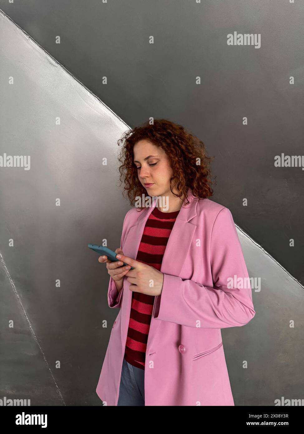 Una giovane donna rossa dai capelli ricci è concentrata sul suo telefono cellulare, rivestita da un elegante blazer rosa su una camicia a righe, con sfondo neutro Foto Stock